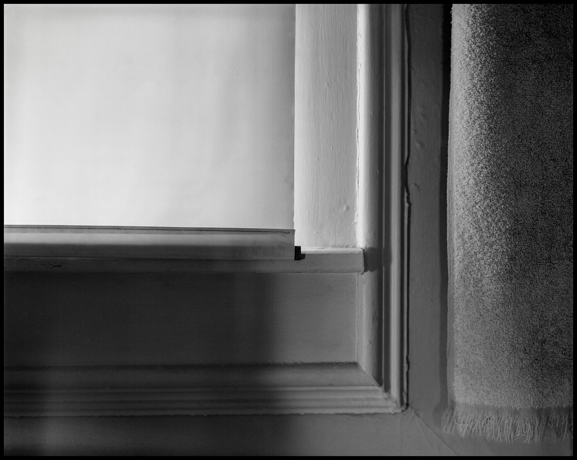 A black and white minimalist abstract study of a bathroom window frame, shade, and towel. Kirksville, MO USA, 1984.