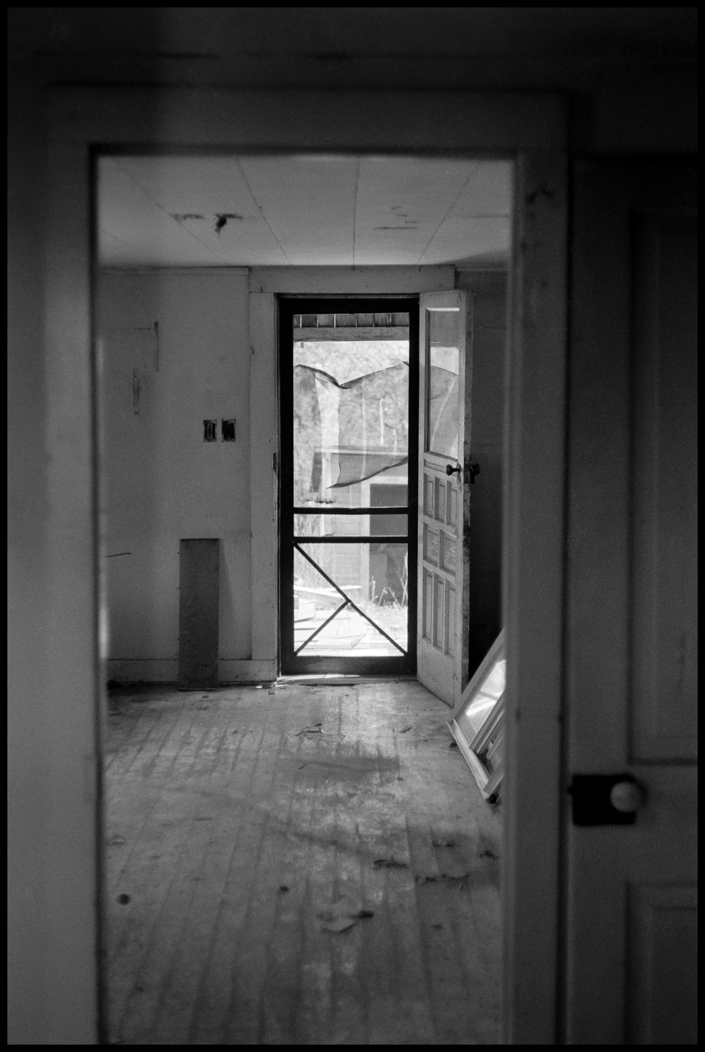 A vintage grainy black and white view from the interior of and abandoned house looking out the back screen door through an interior doorway. Kirksville, Missouri 1977