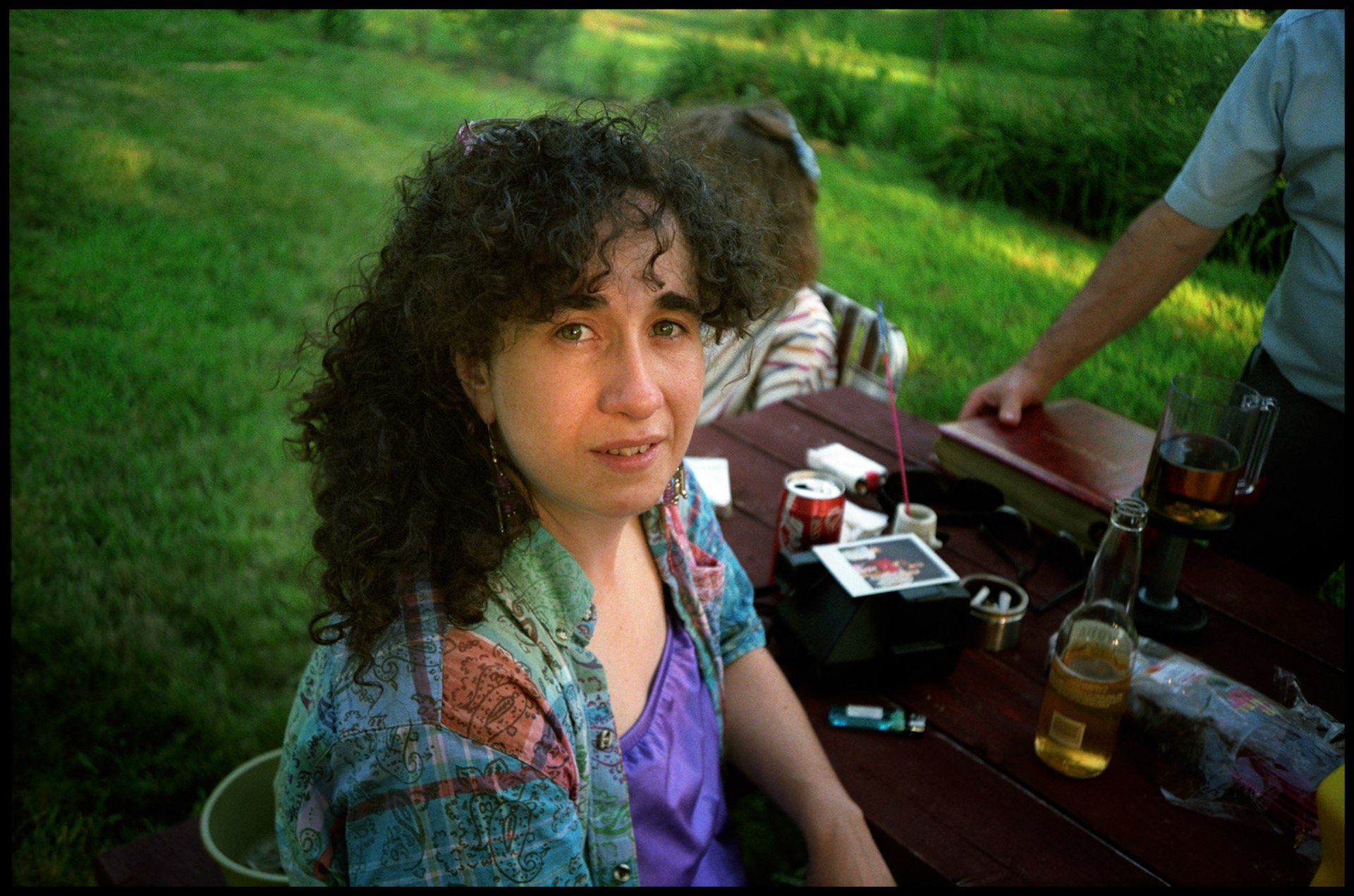 A dark curly haired woman stares inquisitivley at the camera while sitting at a picnic table during an Indepenence Day celebration. Near Kansas City. Missouri 1991