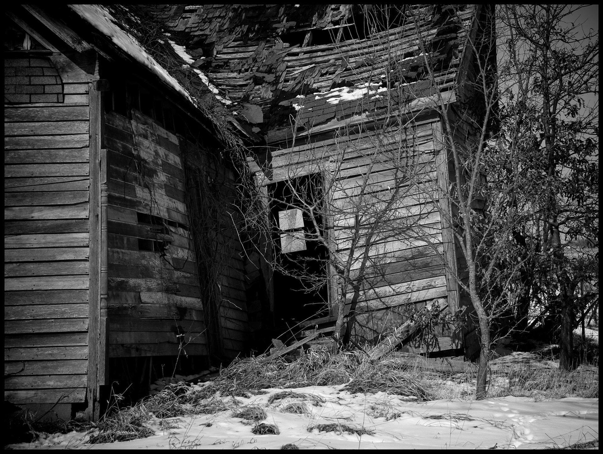 A black-and-white rural Missouri winter landscape detail of a dilapidated abandoned house partially covered in snow looking through an open door to a window on the opposite wall. Near Winigan, Missouri USA 2008.