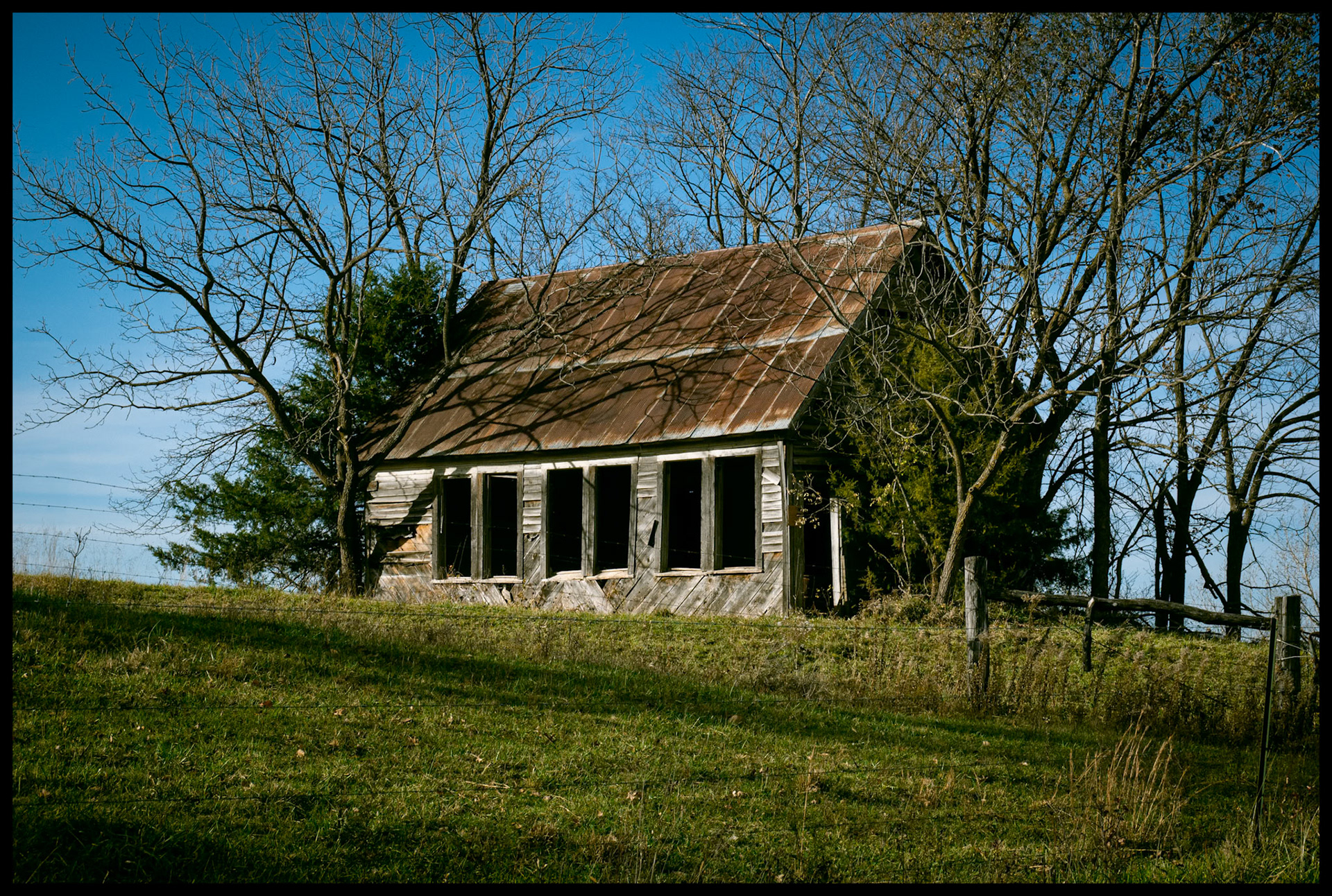An abandoned church building on a green hillside surrounded by mostly bare trees with a whispy clouds in a rich blue sky in the background in late Autumn. Near Ethel, Missouri, 2023.