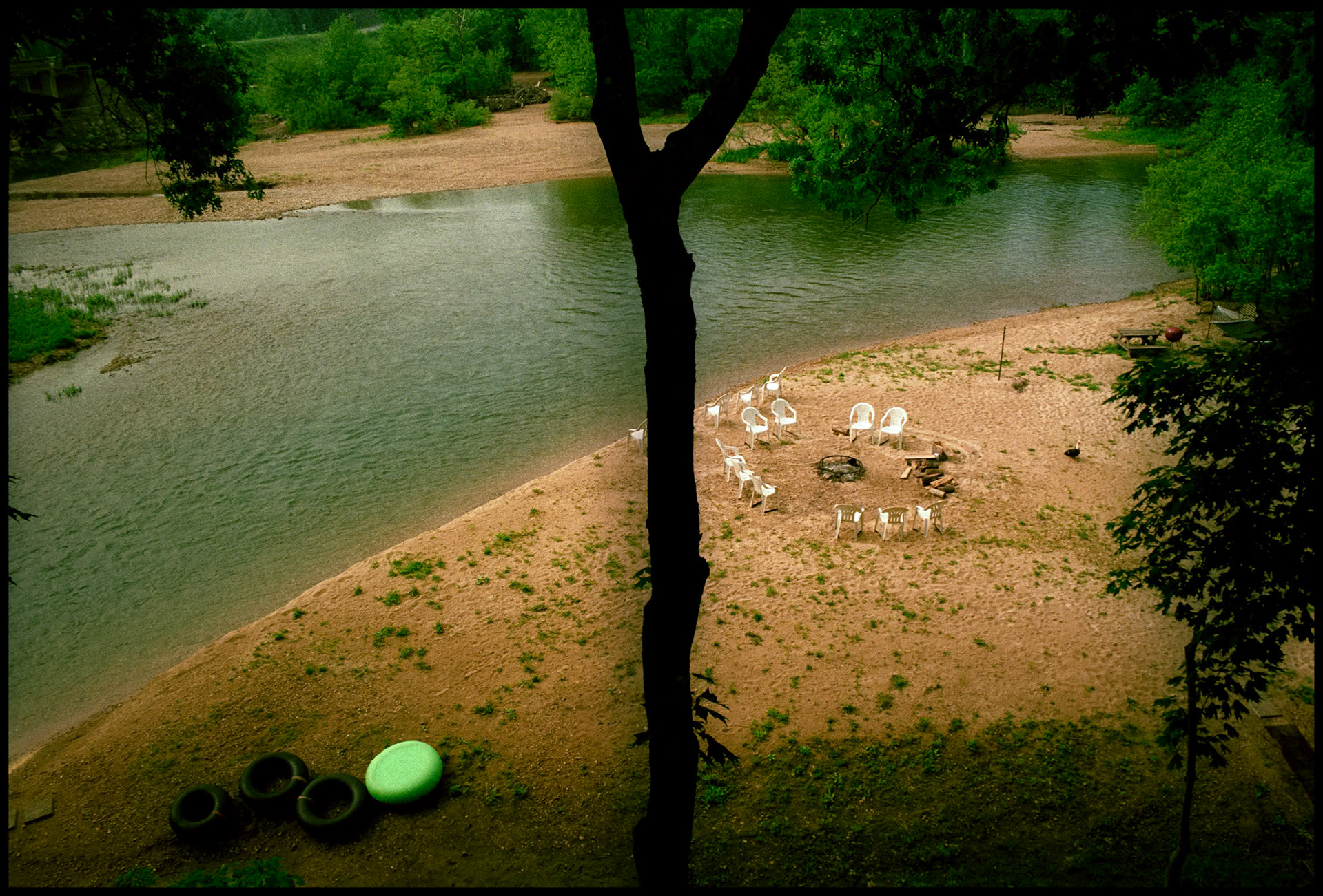 A circle of chairs around a fire pit with inner tubes and a silhouetted tree dividing the view at the edge of the Jack's Fork of the Current River in Eminence Missouri. Shot from the balcony of River's Edge Resort in 1992 and published in the book Images of the Ozarks (University of Missouri Press, 1998)
