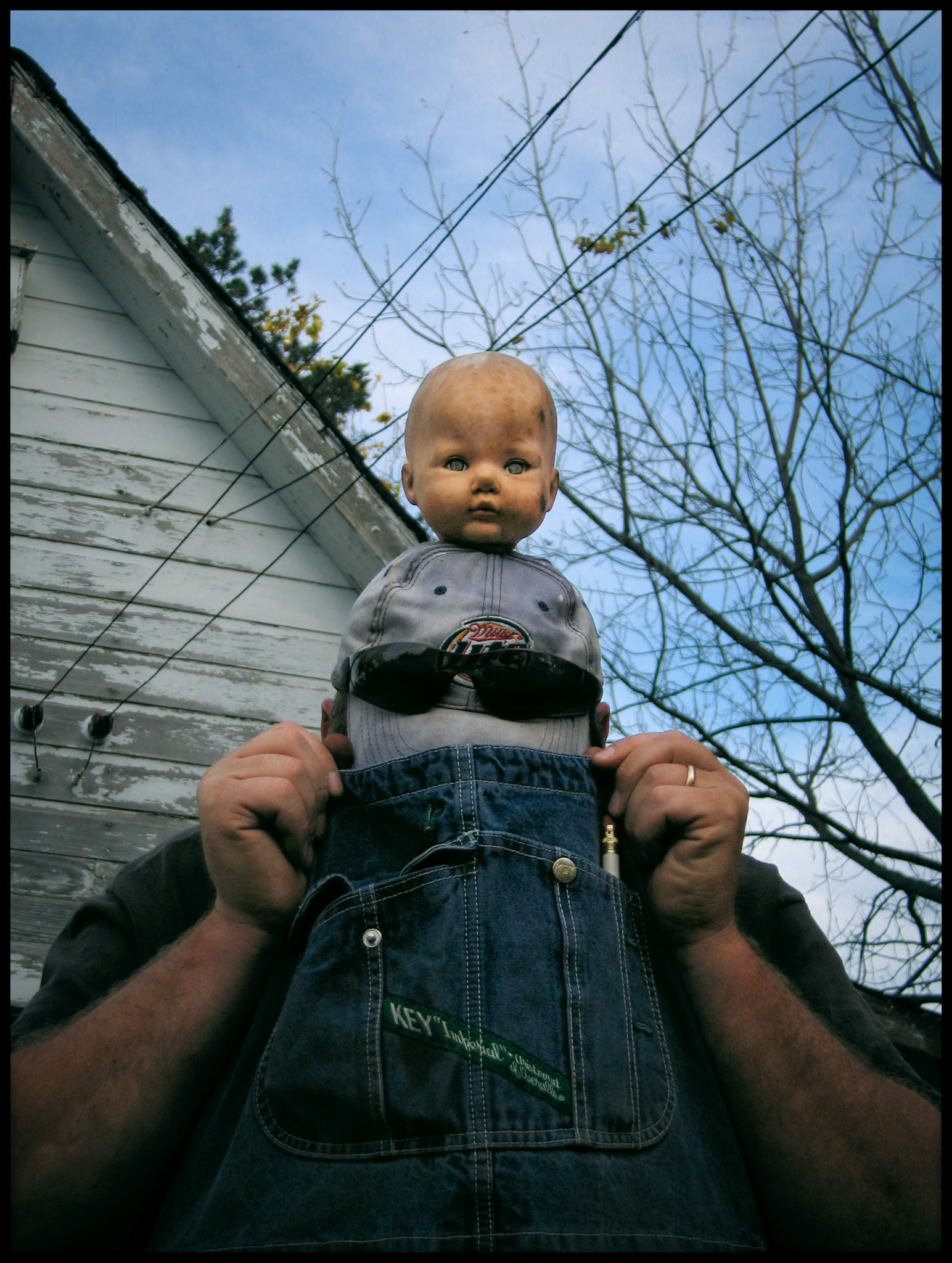 A whimsical, creepy, bizzare image of a man in overalls pulling his bib over his face with a doll head sitting on his cap staring into the lens in front of of wheathered shed. Green City, Missouri 2006