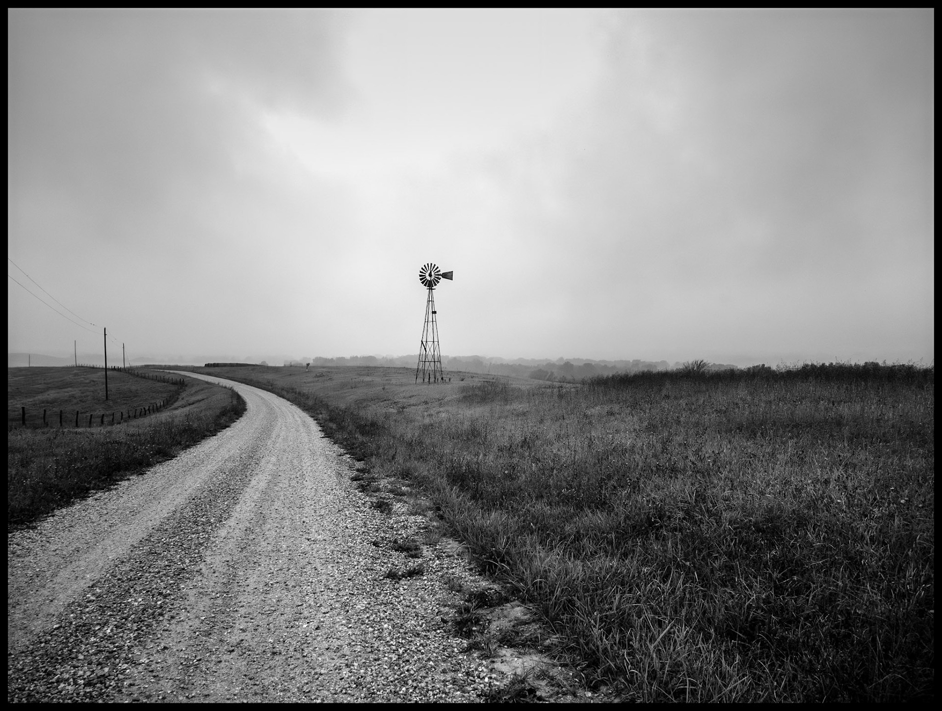A solitary windmill at the edge of a gravel road and green field silhoutted against the overcast sky on a rainy and foggy September morning. Located on Indian Hill near South Gifford, Missouri, 2023.