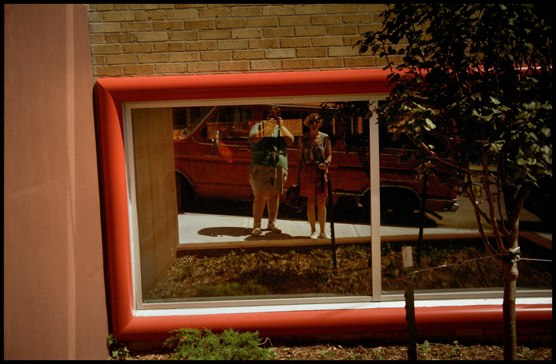 Colorful abstract architectural detail of red framed window with the reflection of the male photographer and his woman friend who is holding her camera. Kansas City, Missouri, 1990