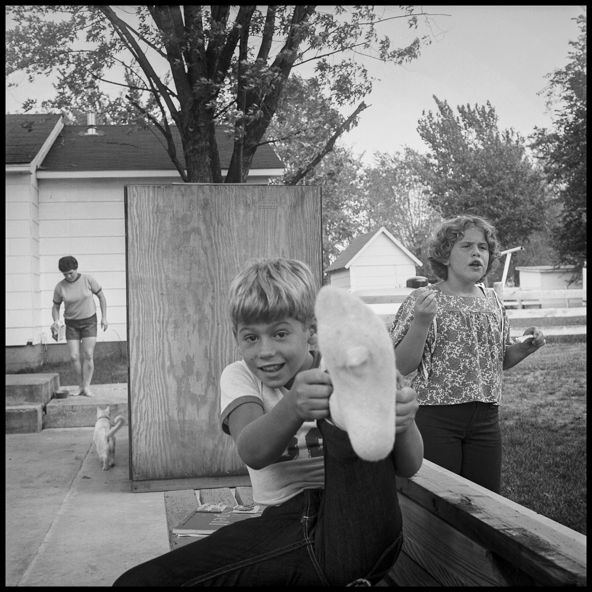 Vintage image of young boy holding his foot up to reveal his dirty sock with his sister, mother, and pet cat in the background on their patio. Kirksville, Missouri 1977