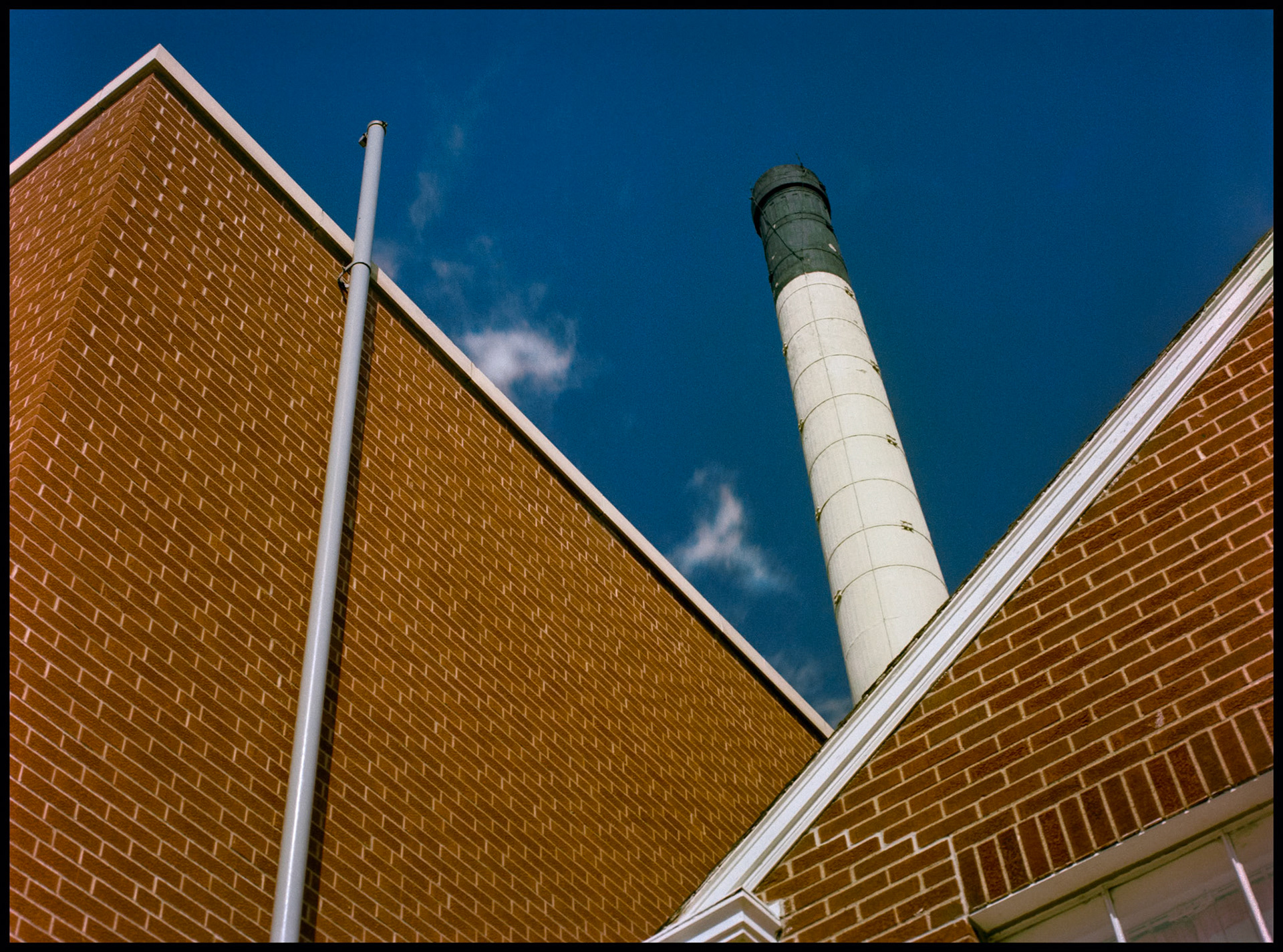 A minimal abstract detail of a pipe and smokestack framed by the rooflines and brick wall on the Northeast Missouri State University (Since renamed Truman State University) Physical Plant building. Kirksville, Missouri 1981