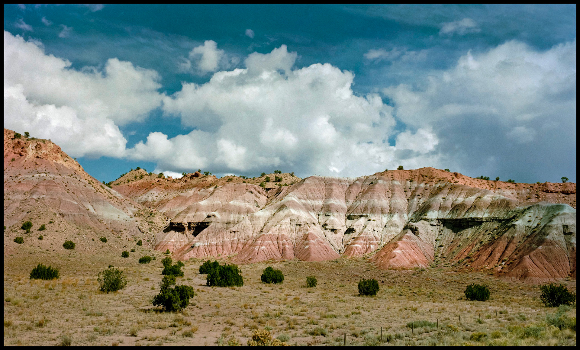 A hillside in New Mexico with muti-colored striped layers and a deep blue sky festooned with billowing white clouds. New Mexico USA 1993