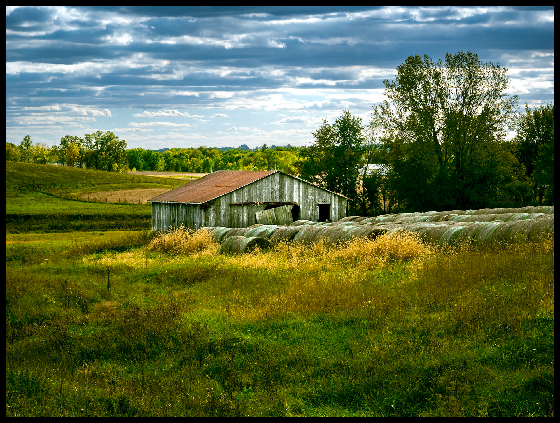 Large bales of hay lined up in front of an old barn surrounded by grass and trees beginning their Autumn color change against a scattering of dark and light clouds at the base of Indian Hill near South Gifford, Missouri. 2023