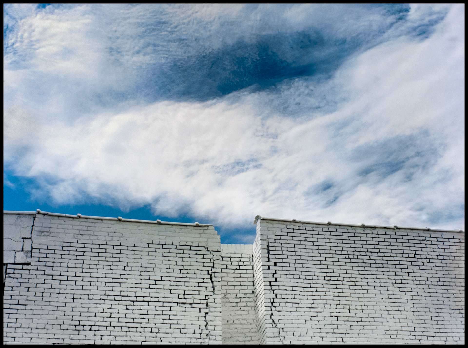 A minimal abstract detail of a white brick wall of a commercial building, puffy white clouds against a blue sky with a power line crossing it in the industrial area of the West Bottoms in Kansas City, Missouri. 1984
