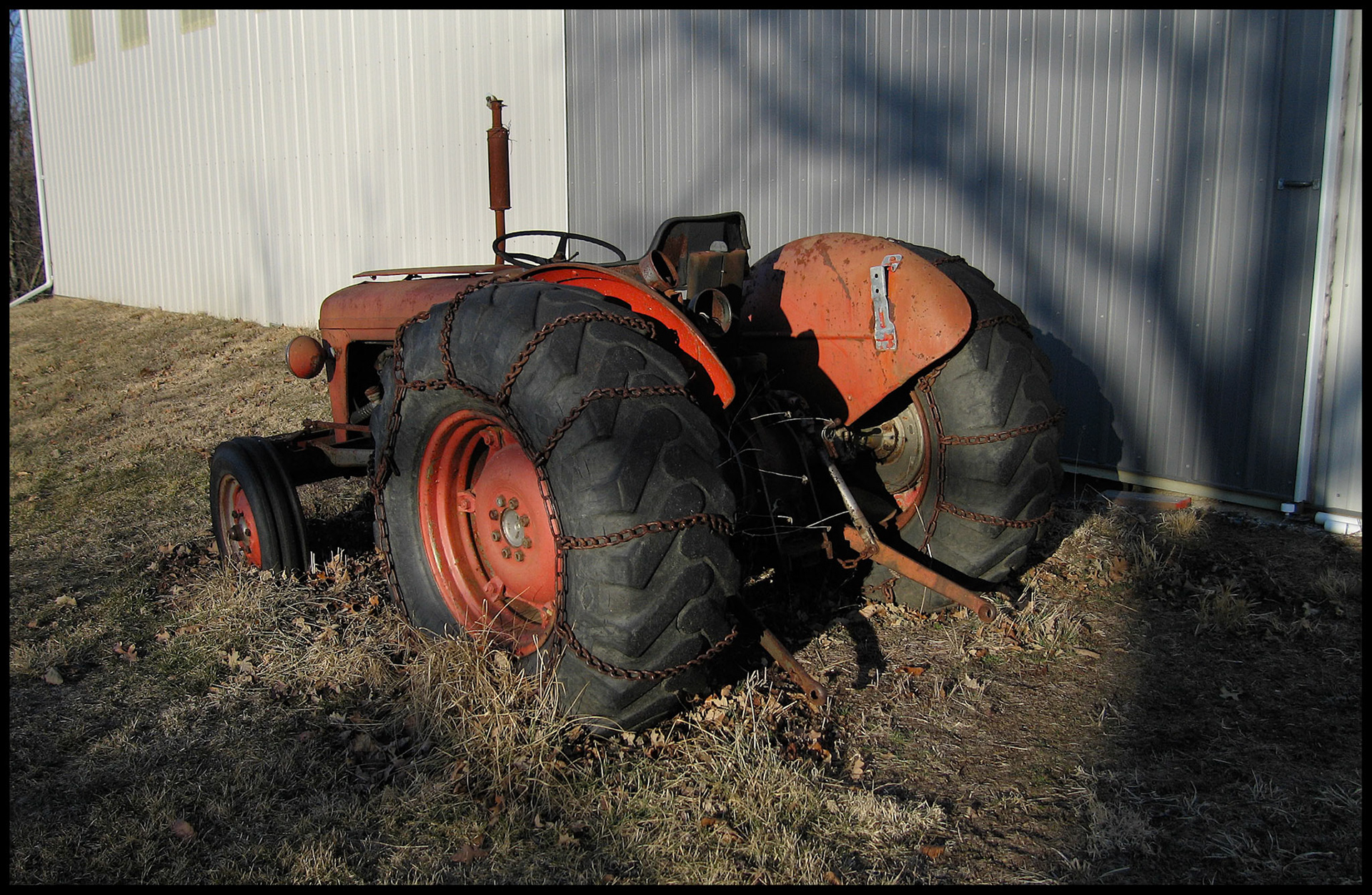 Orange vintage tractor next to metal building with snow chains on rear tires highlighted by late afternoon sunlight.