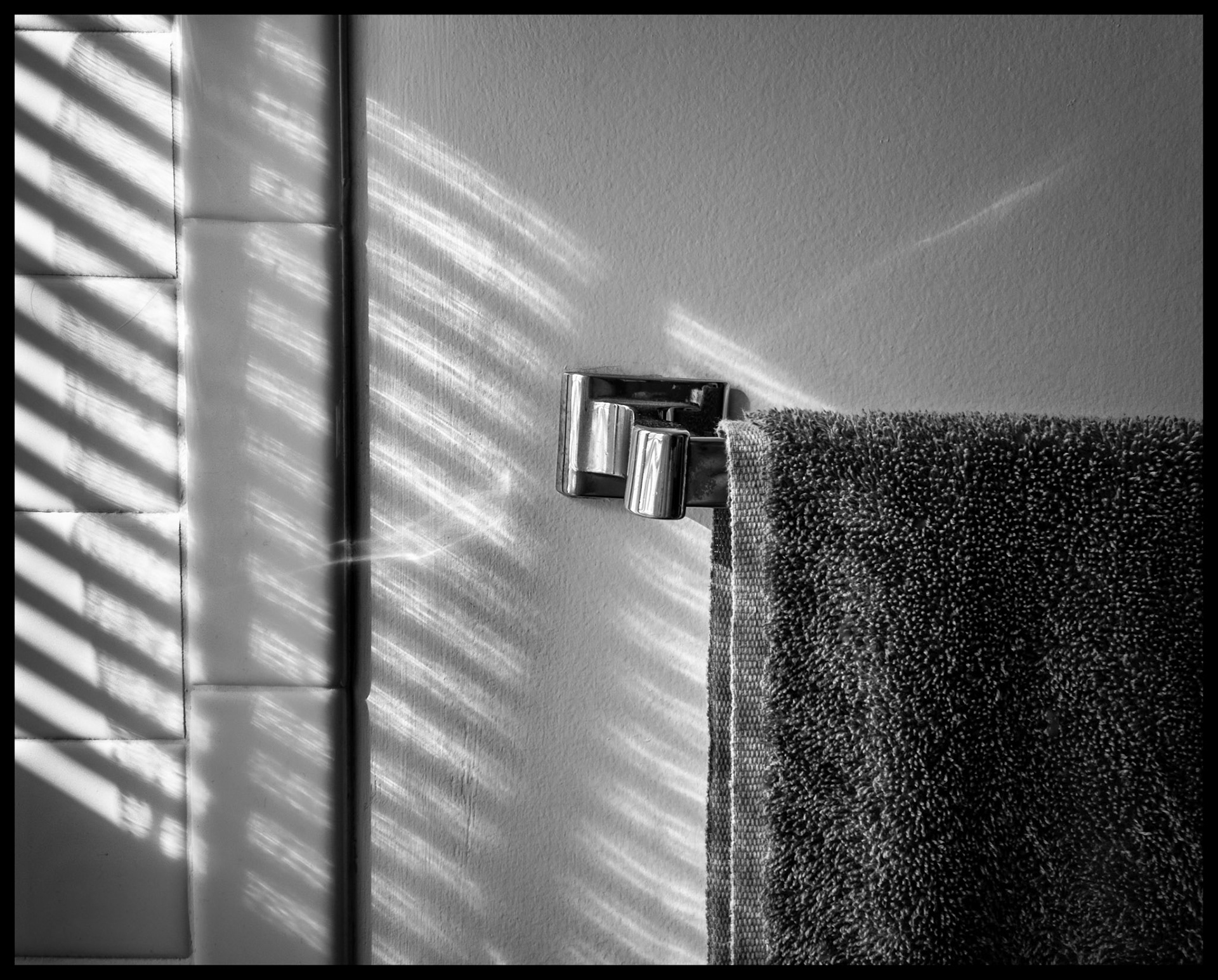 A minimal abstract still life view of highlights and shadows from a window blind on a bathroom wall and towel rack. Kirksville, Missouri 2022