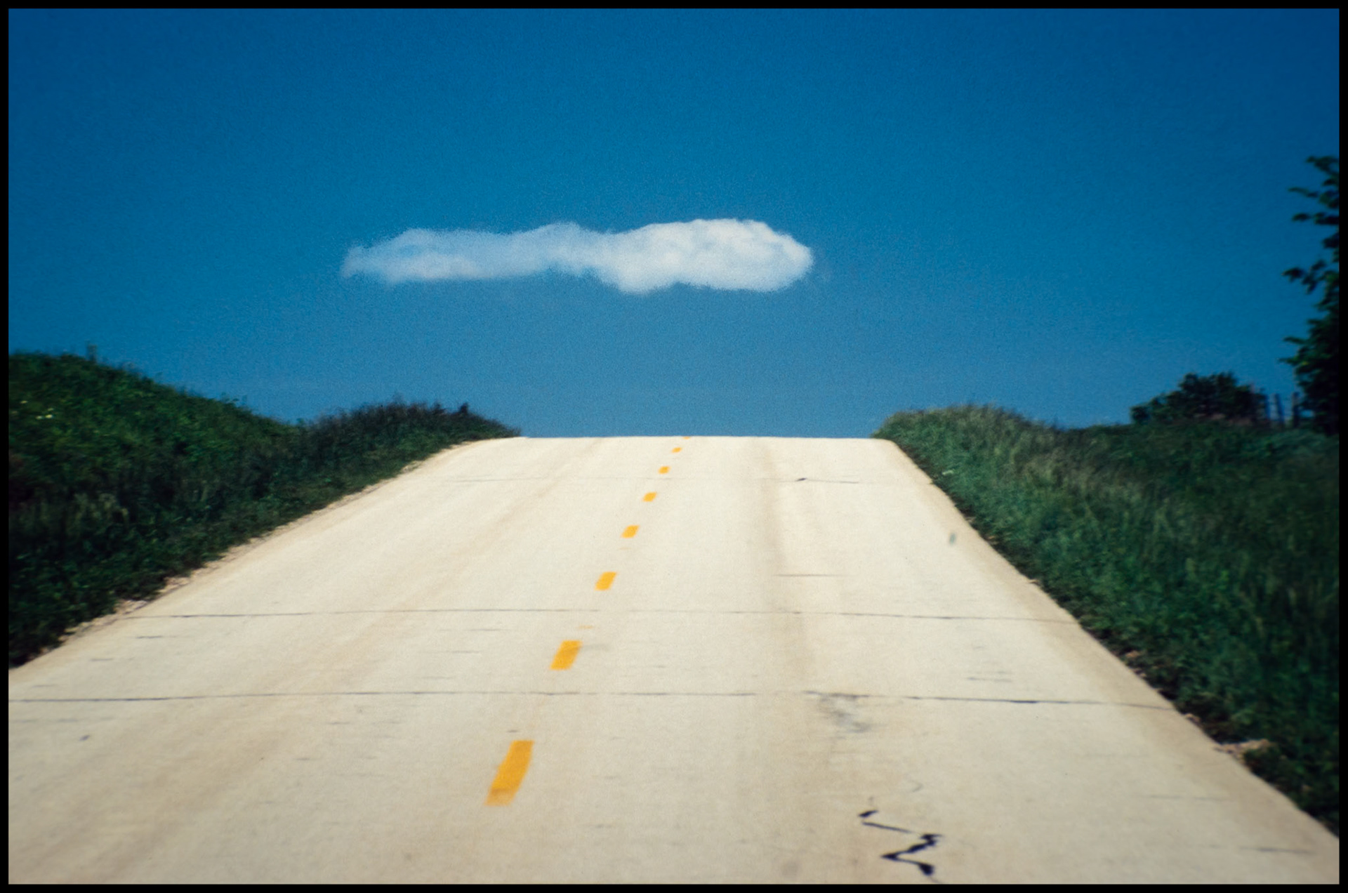 A grainy image from the 1980s of an old bright cement highway leading up a steep hill, seemingly, toward a tadpole shaped cloud at the top.