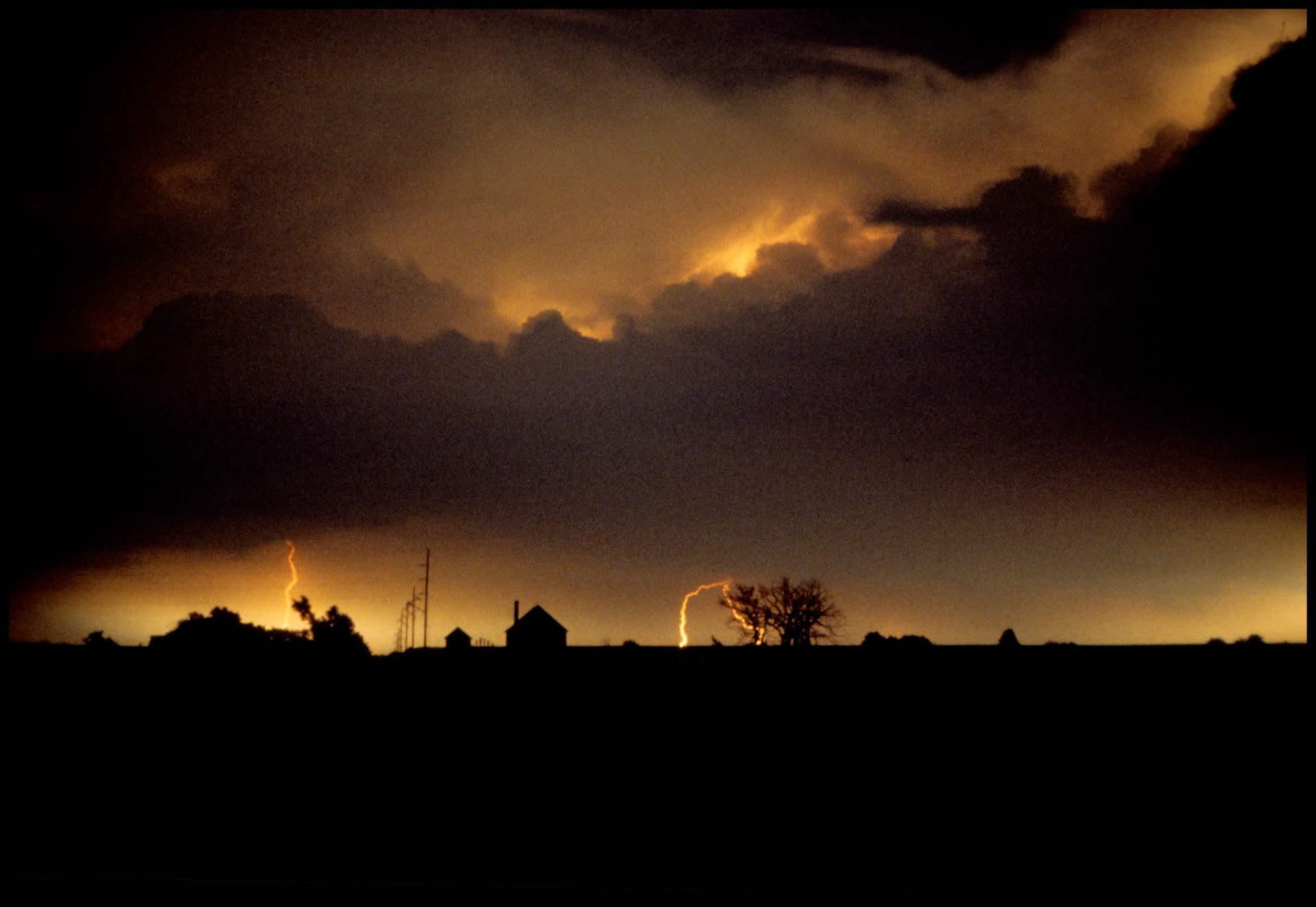 A grainy time exposure night shot of a thunderstorm lighting up the sky and lightning bolts striking the horizon in two places leaving the two houses and row of power poles in silhouette.  Near Brashear, Missouri, 1980.