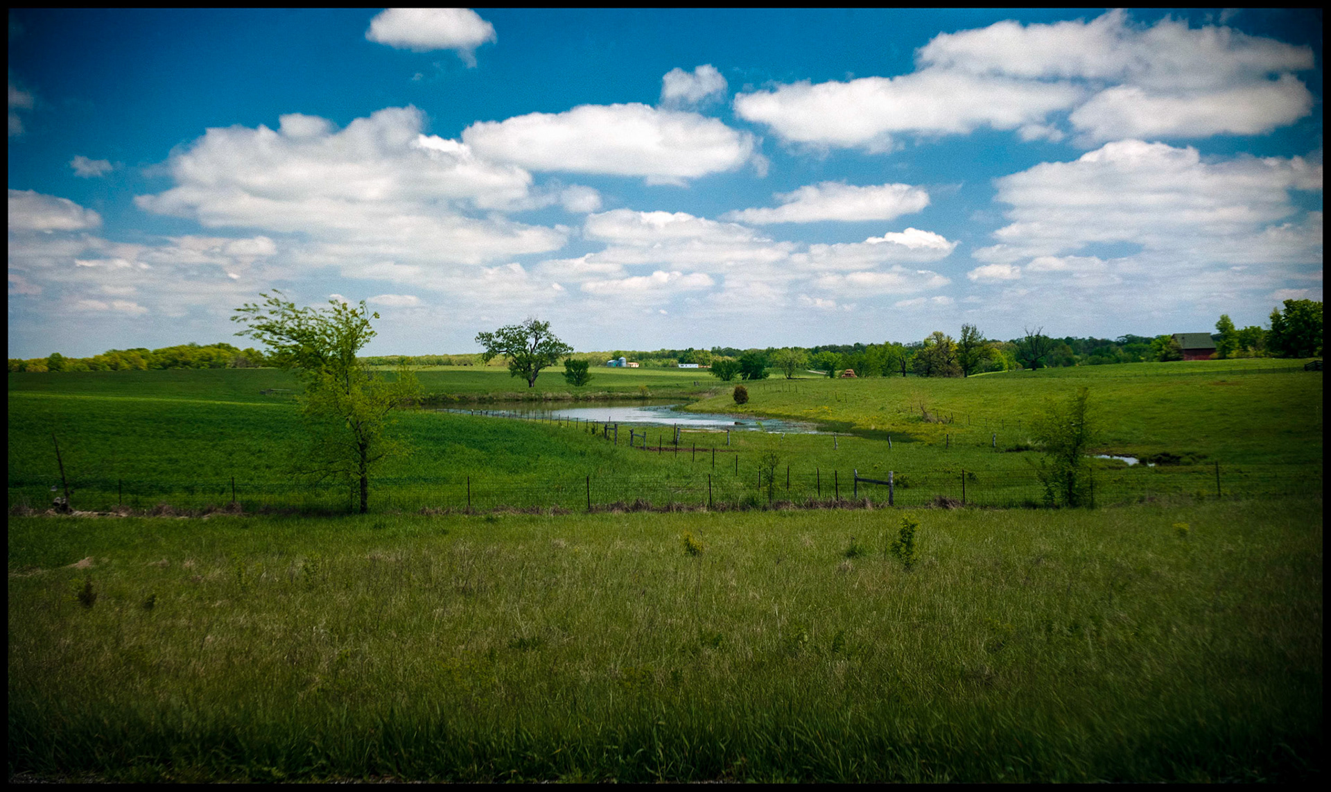 Green meadow and waterway with a farm house and buildings highlighted by the sun through a break in the puffy clouds making up the background, near Macon, Missouri 1990