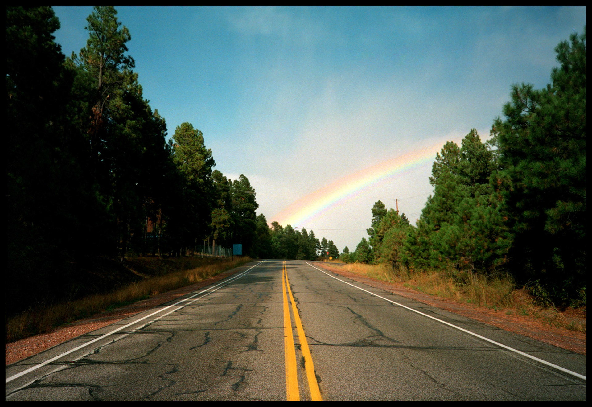 The double yellow lines of a lonely Highway in New Mexico seemingly leading to the base of a rainbow; evoking idealism, optimism, and a sense of a reward at the end of your journey. Near Los Alamos New Mexico USA 1993