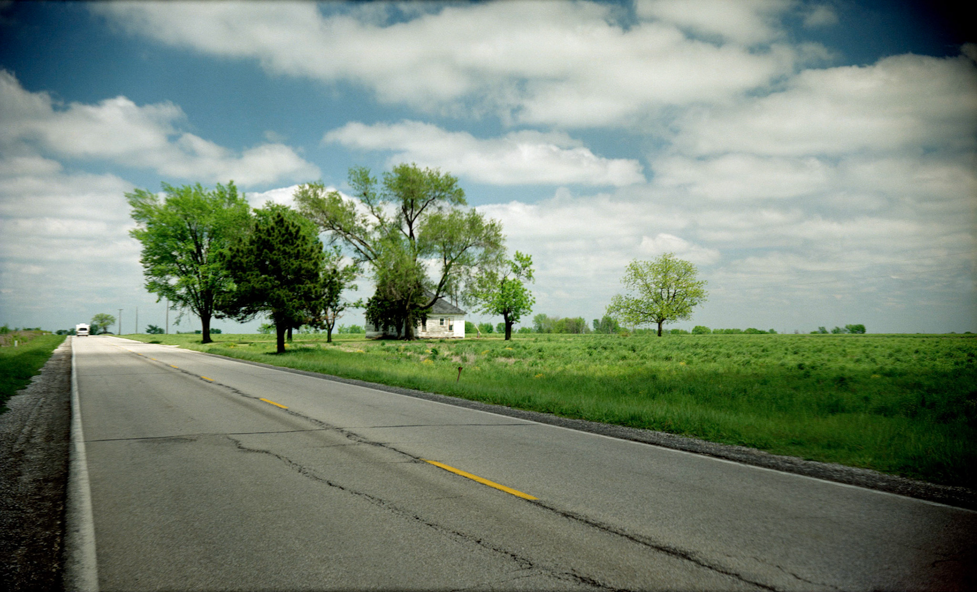 Soft focus image shot with an antique camera of two vehicles highlighted by the sun heading on down a rural Missouri two lane highway next to an abandoned house with puffy clouds in the background.
