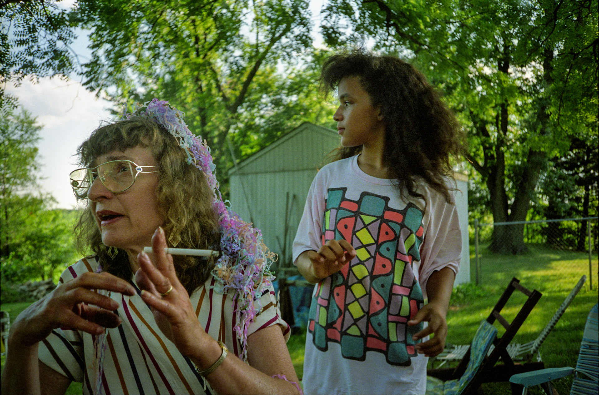 Older woman with cigarette in her hand wearing a headdress made from popper firework streamers on the 4th of July in her back yard as her teenage granddaughter looks on. Near Kansas City, Missouri 1991