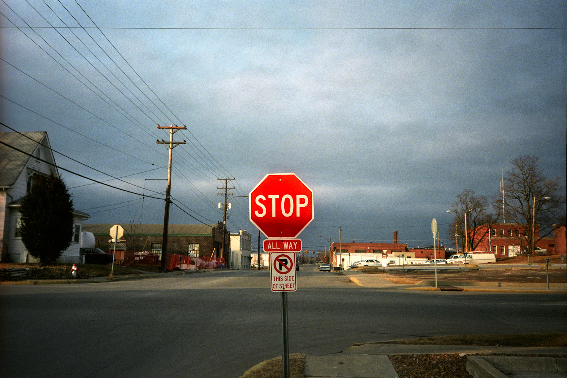 An urbanscape with a sunlit bright red stop sign as the center of attention at the intersection of an industrial area of Columbia Missouri in 1994 with purplish gradated clouds in the background and a row of power lines and poles to the side. Columbia, Missouri 1994