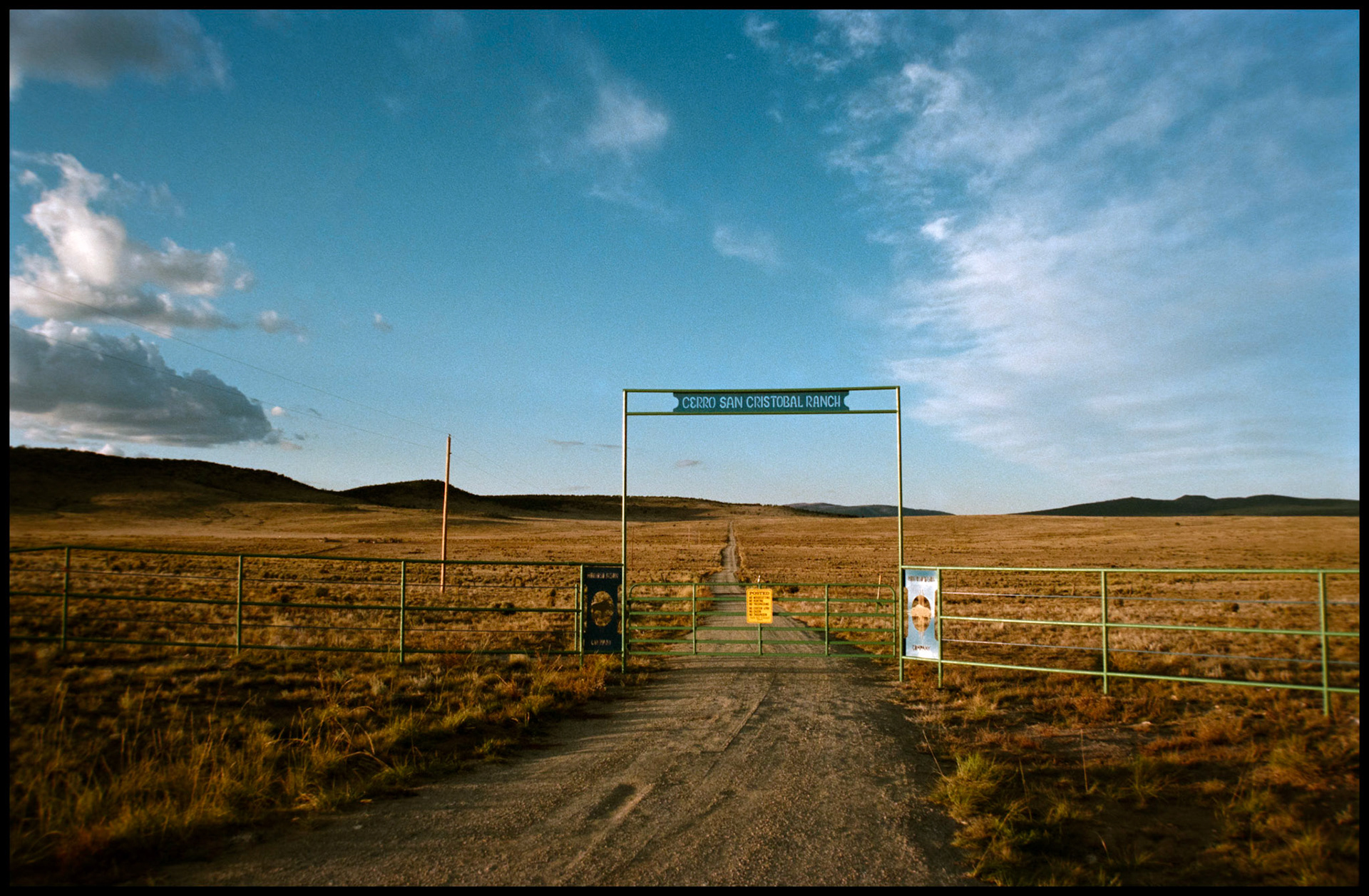 The gate of a ranch with a road leading off toward the horizon and silvery clouds adorning the sky. Near Taos, New Mexico USA 1993