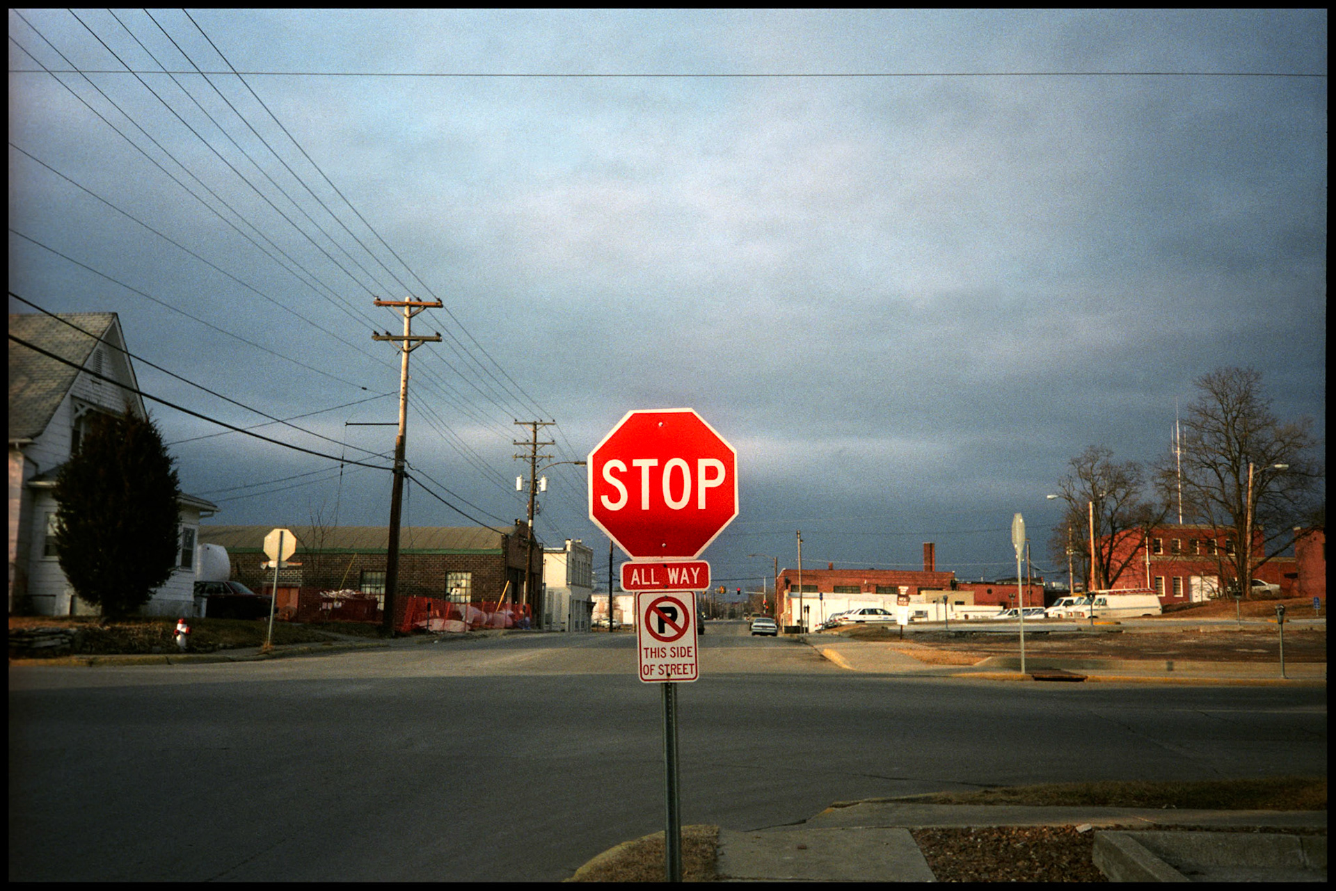 An urbanscape with a sunlit bright red stop sign as the center of attention at the intersection of an industrial area of Columbia Missouri in 1994 with purplish gradated clouds in the background and a row of power lines and poles to the side. Columbia, Missouri 1994