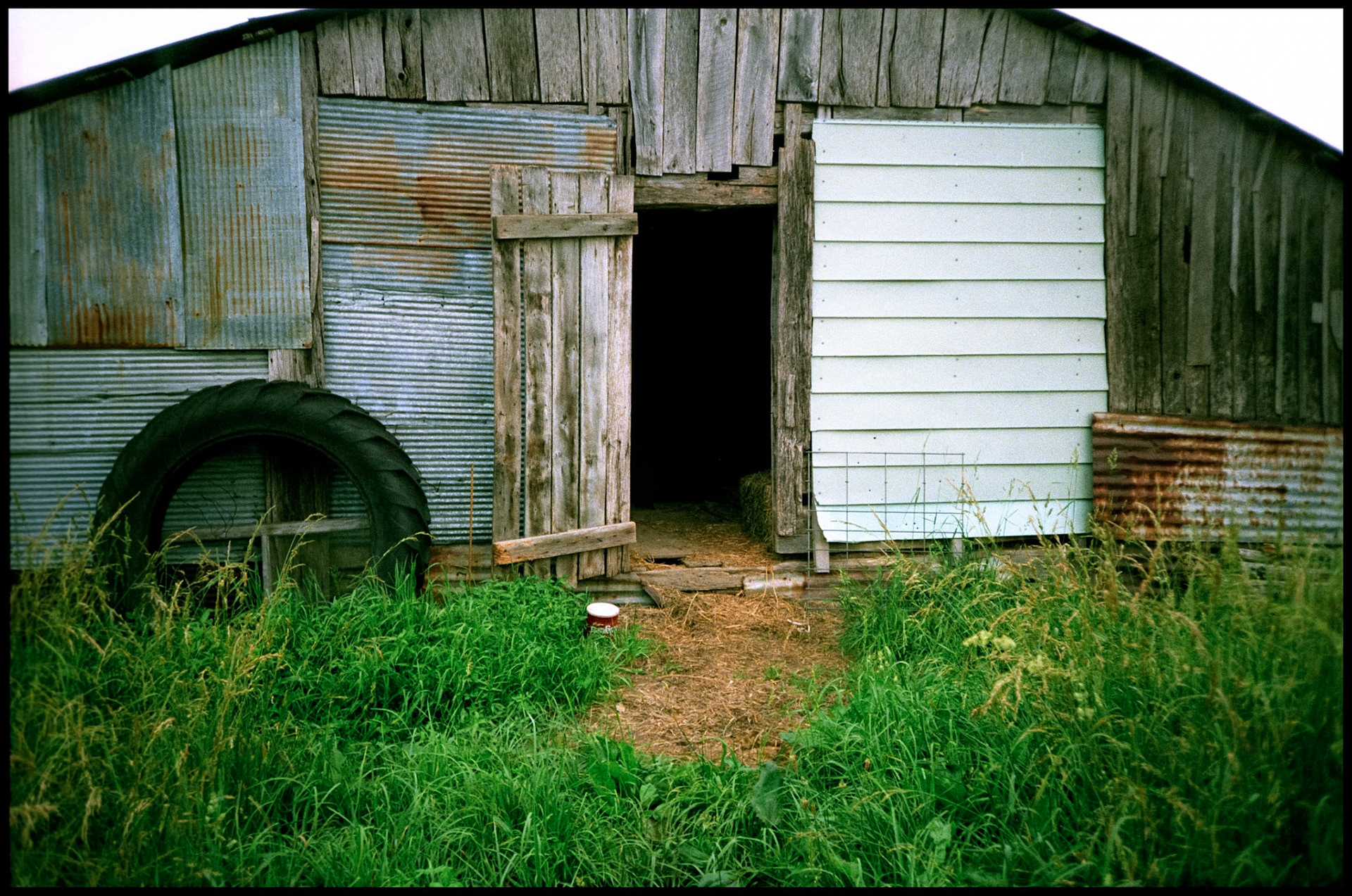 A weathered wooden farm building patched with corrugated metal roofing material and vinyl siding with an old tractor tire leaned against the front near Harrisburg, Missouri 1991.