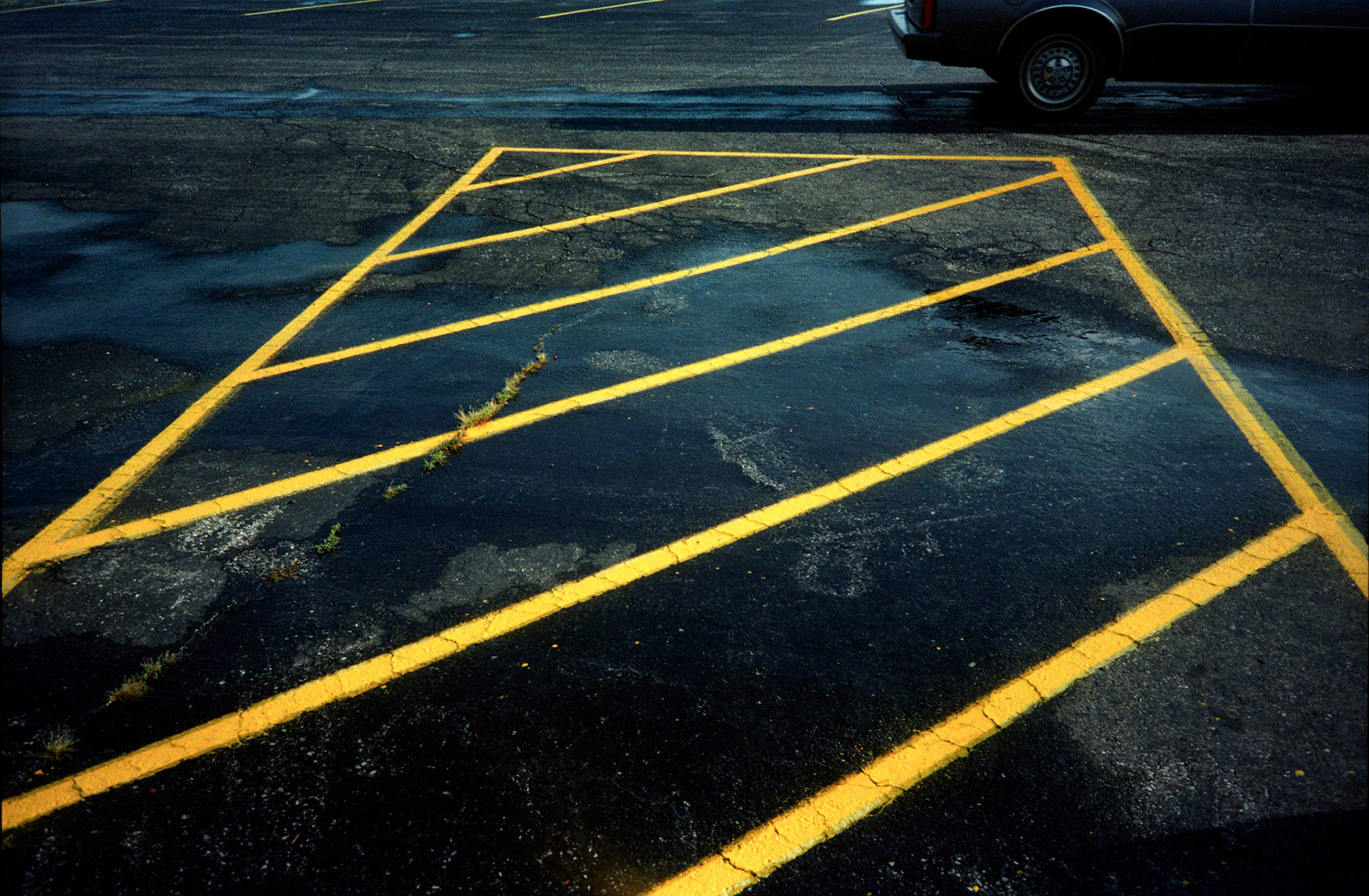 An minimal abstract detail of bright yellow diagonal lines in a black asphalt parking lot making an interesting design with the rear tire of a car in the background. Centralia, Missouri 1994