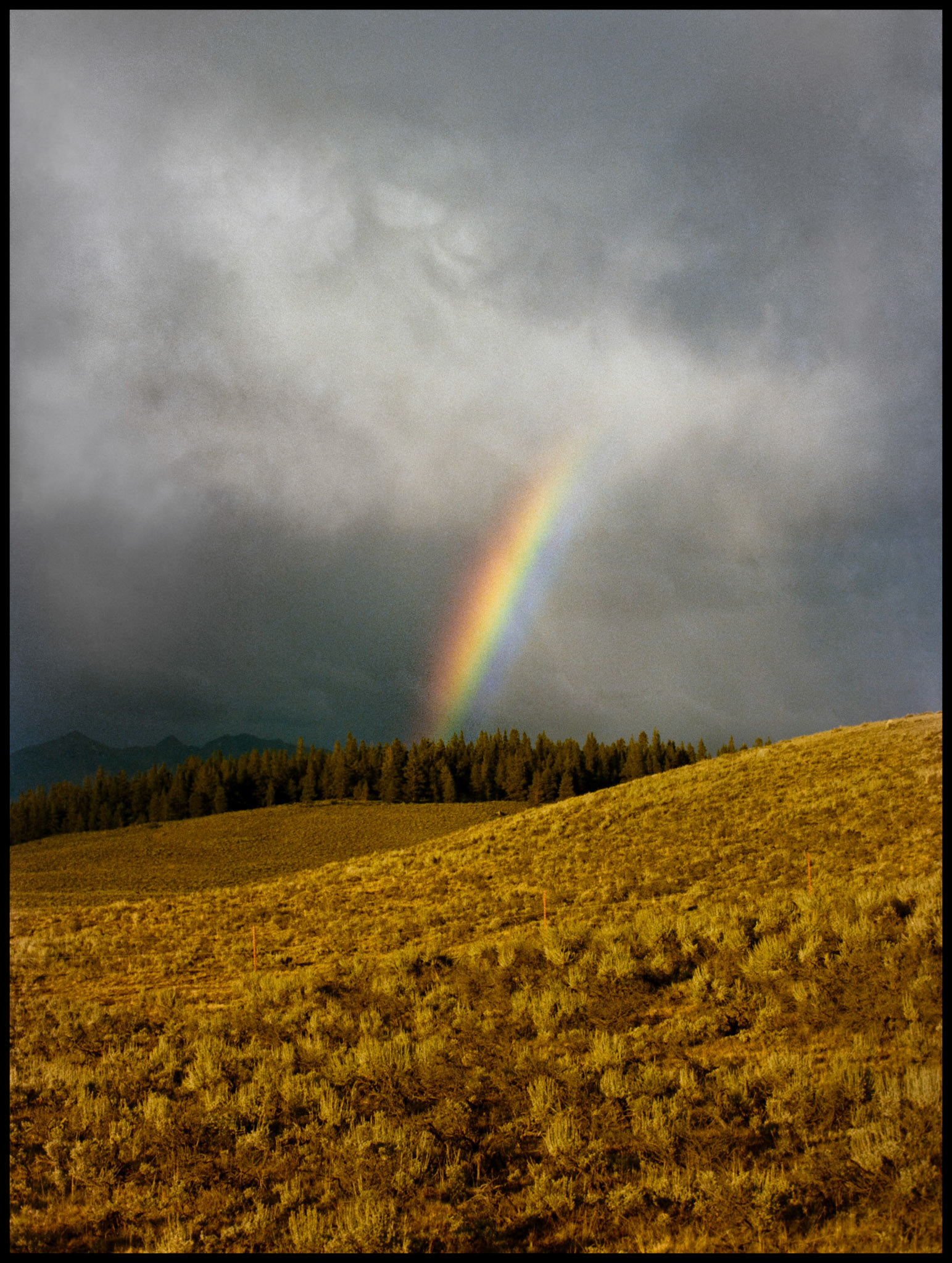 A bright colorful rainbow highlighted by the afternoon sun ahead of the storm contrasted by the swirling dark gray rain clouds coming down from the mountains in the background. Taylor Park near Tin Cup, Colorado 1978