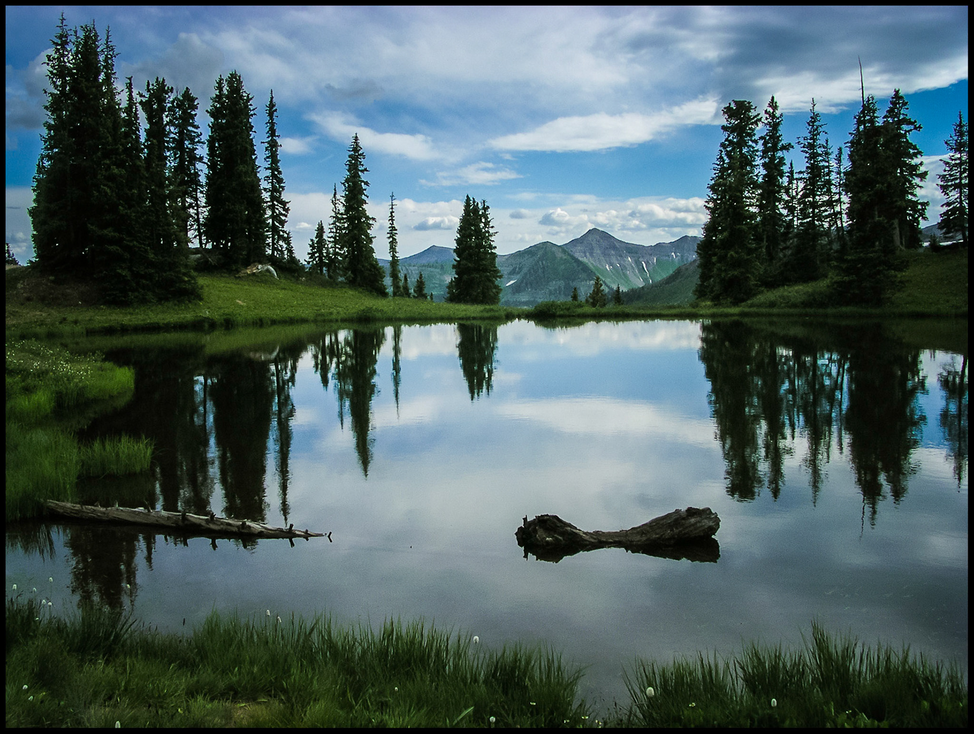 A serene view of an Alpine lake reflecting the blue sky, clouds, and surrounding pine trees with mounain tops at eye level in the distance. Paradise Divide near Gothic, Colorado USA 2004.