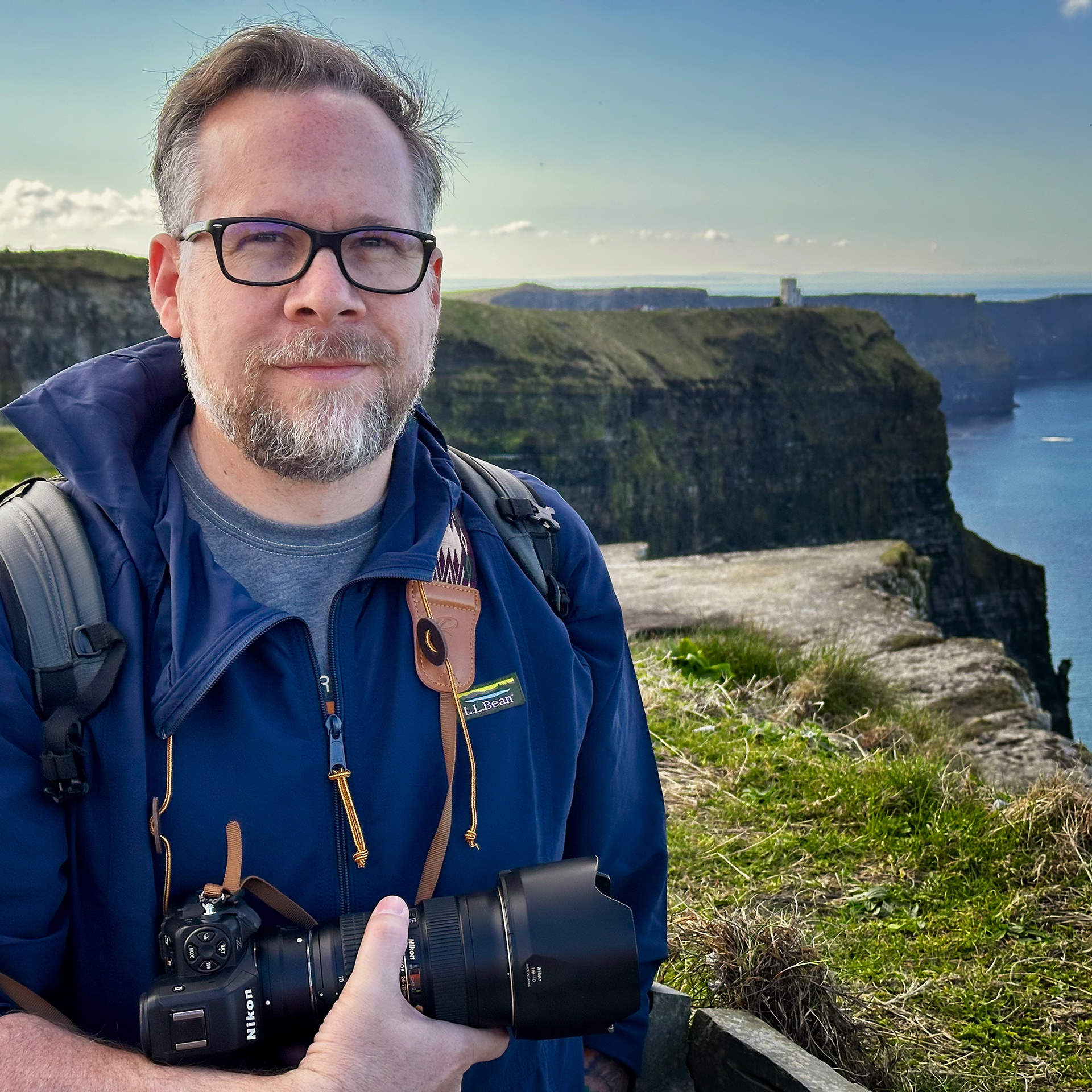 Man with glasses holding a professional camera, standing near the Cliffs of Moher in Ireland with the ocean and cliffs in the background.
