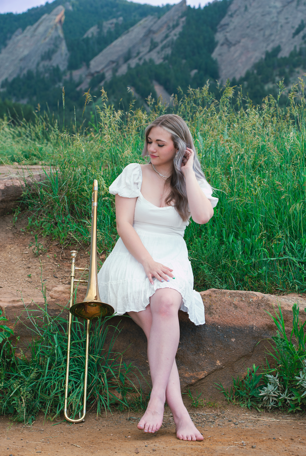 Boulder senior portrait of a high school girl in a white dress, sitting barefoot on a rock with the Flatirons in the background and a trombone resting beside her.