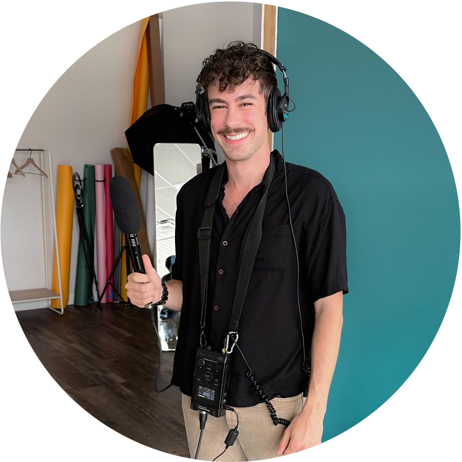 A man with brown curly hair and a mustache wears a black button-up short sleeve shirt and brown pants. He is smiling and holding a microphone with headphones on.