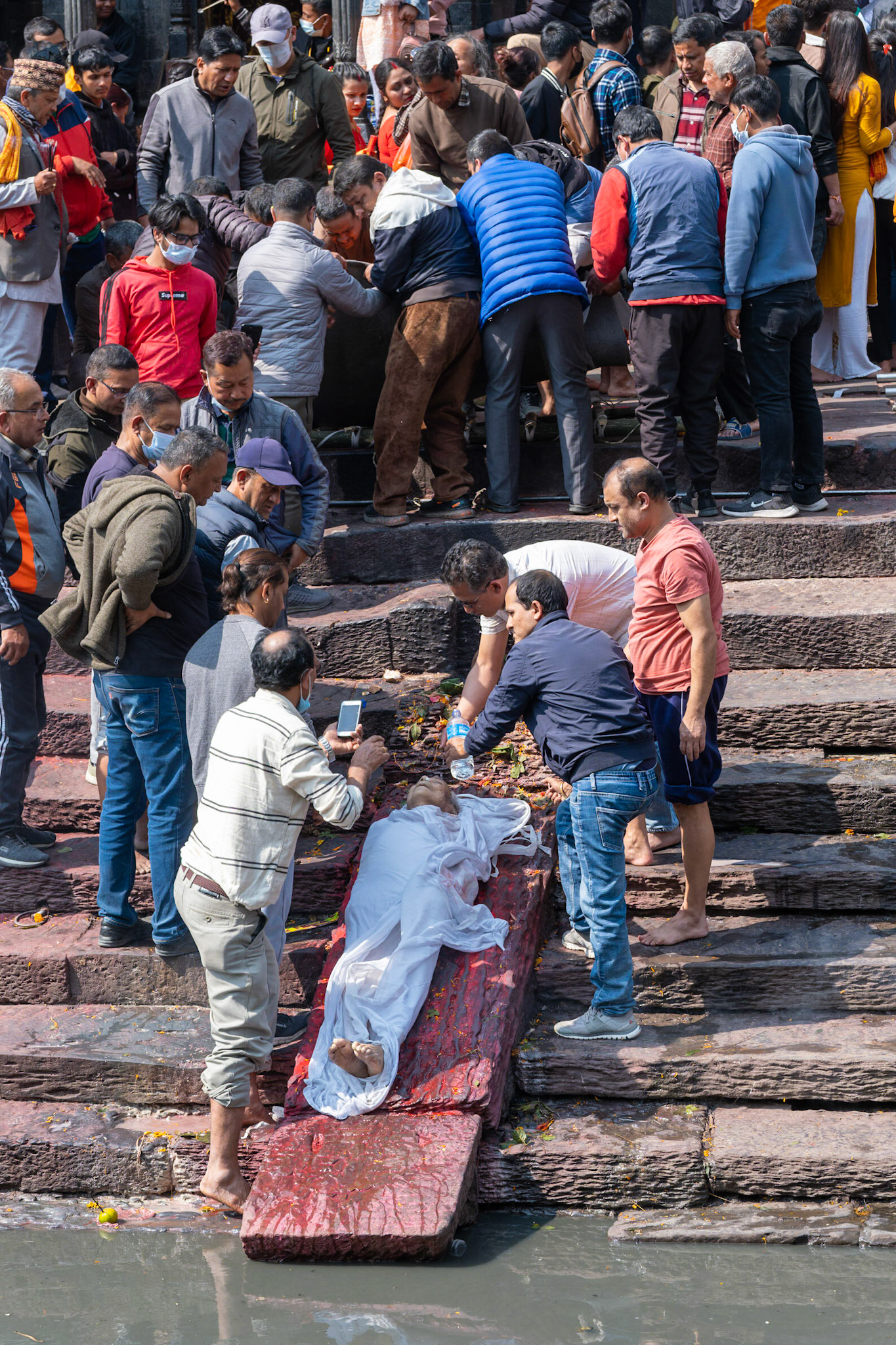 Une famille lave symboliquement sa défunte avec l'eau sacrée de la rivière Bagmati qui se jette dans le Gange. A gauche, un homme prend une photo du corps. en amont, un groupe d'homme installe un autre corps sur une civière afin de l'installer sur la rampe lorsque la vielle dame sera partie de l'autre côté du pont pour la crémation. Je me trouve de l'autre côté de la rivière, avec des milliers d'autres personnes. Certaines sont indifférentes à la scène, d'autres, comme moi, sont fascinées par ce spectacle qui peut nous sembler d'un autre temps, mais qui est tout simplement d'une autre culture.