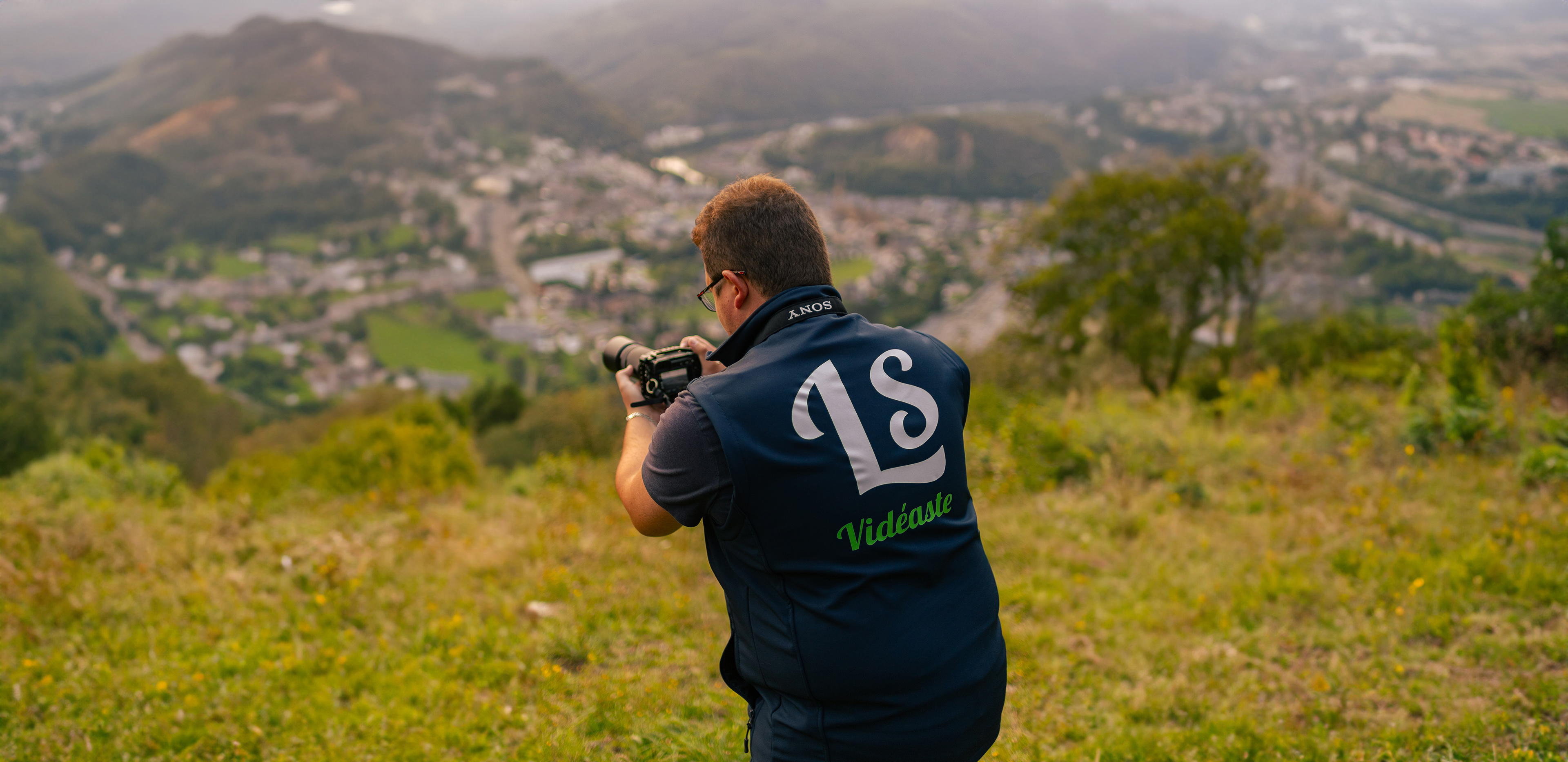 Photographie de Leroy Steven prise de dos alors qu'il capture l'incroyable beauté de la vallée de la Meuse dans les Ardennes. La vallée se dessine en arrière-plan, créant un magnifique effet de flou qui met en valeur la scène capturée.