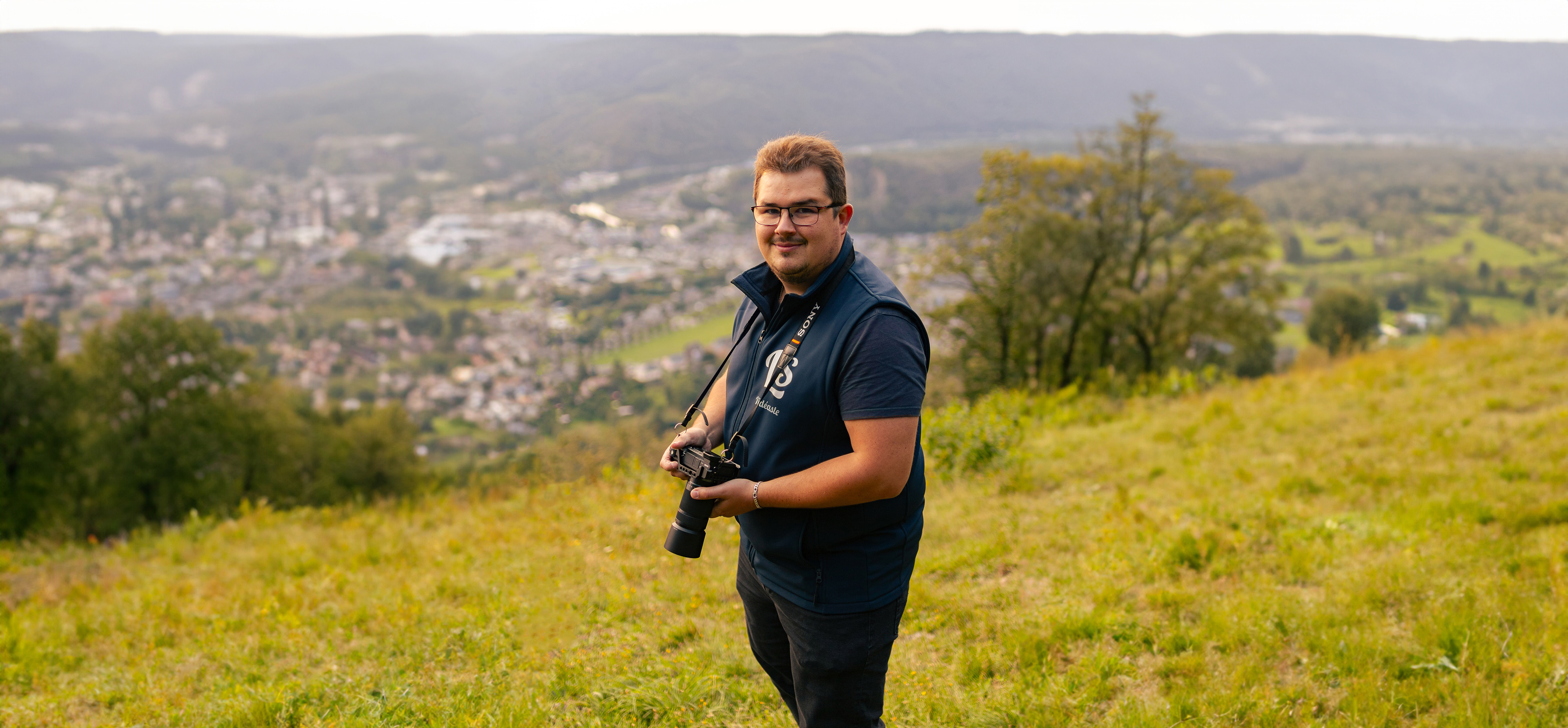 Photographie de Leroy Steven tenant un appareil photo au sommet d'une colline, avec une vue panoramique sur la vallée de la Meuse dans les Ardennes. La composition met en valeur un magnifique flou d'arrière-plan, créant une atmosphère saisissante.