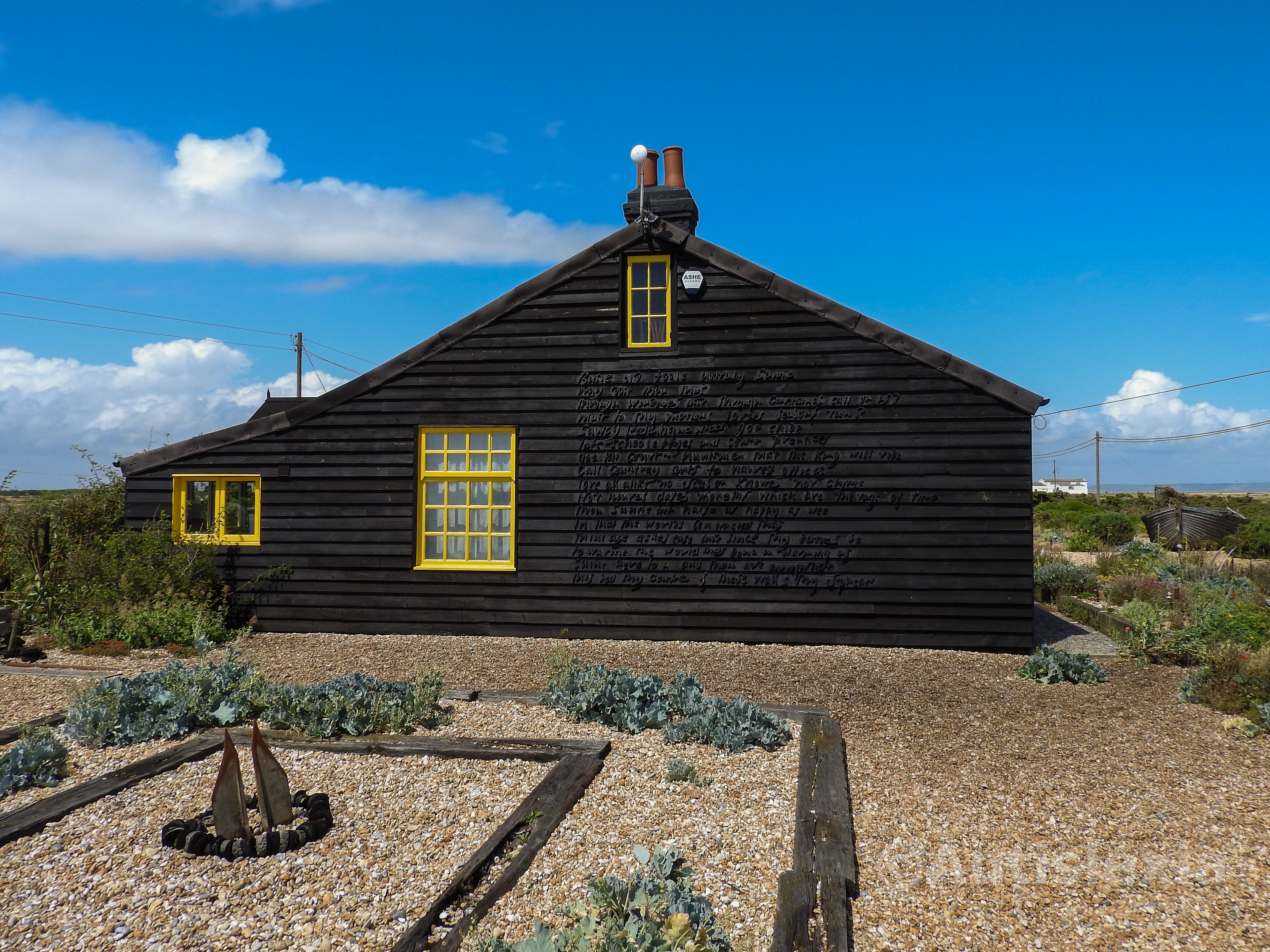 Image of the side of Prospect Cottage (by Derek Jarman) ; dark wooden cottage with bright yellow window frames sitting on gravel with its landscaped garden around it.