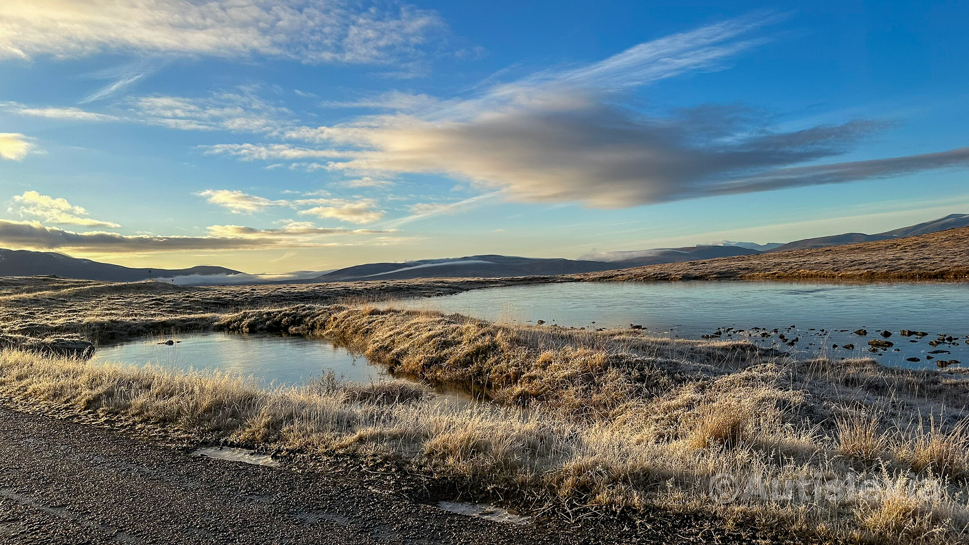 Frosty scenery with the edge of a road in the bottom left, and grass and water beyond with mountains in the background and a blue sky with wispy white clouds