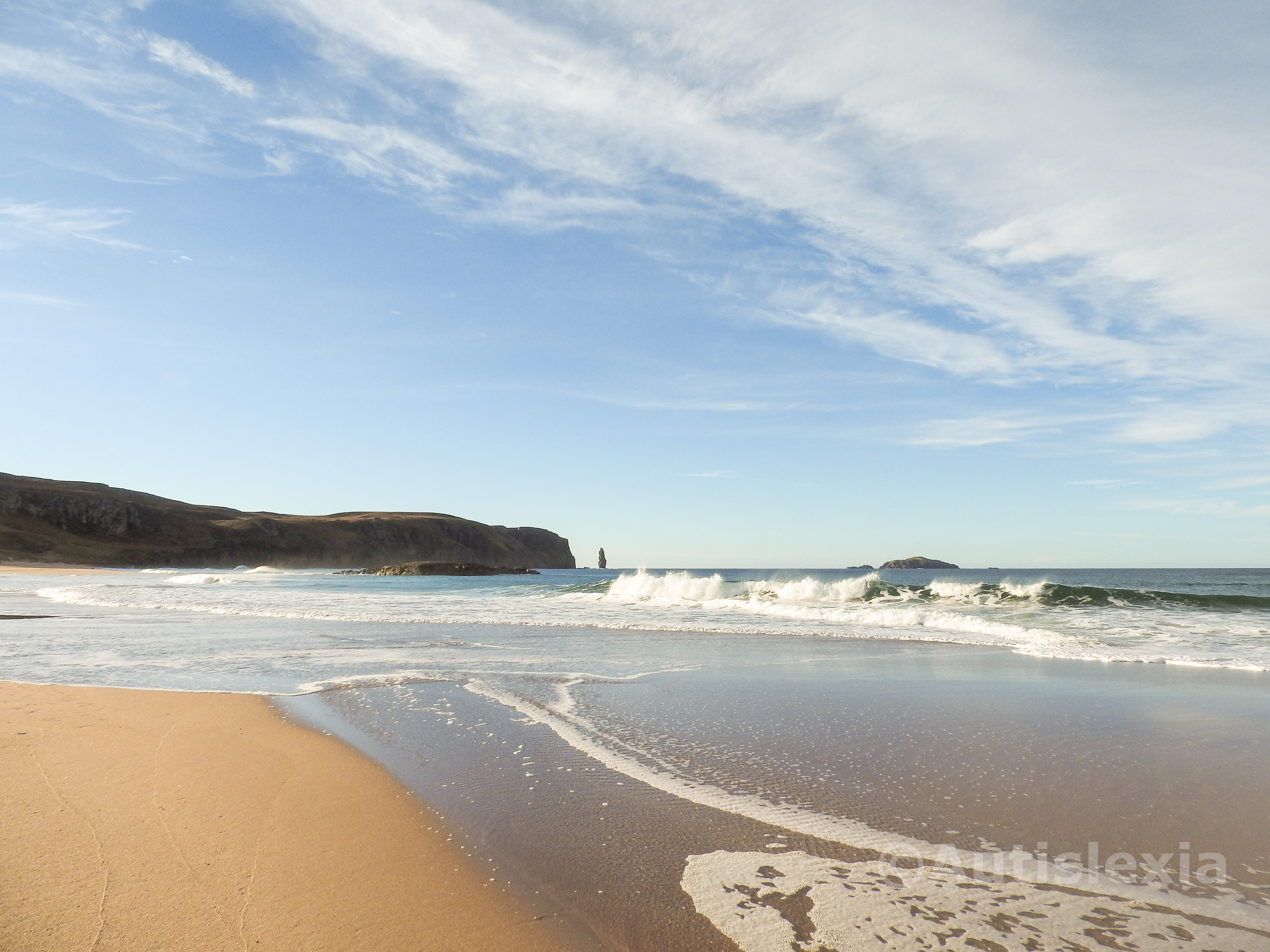 Image taken standing on the Sandwood Bay beach, with the waves breaking and running up the beach. There are the cliffs in the distance on the left with the stack at the end of the cliffs and a blue and white cloud sky.