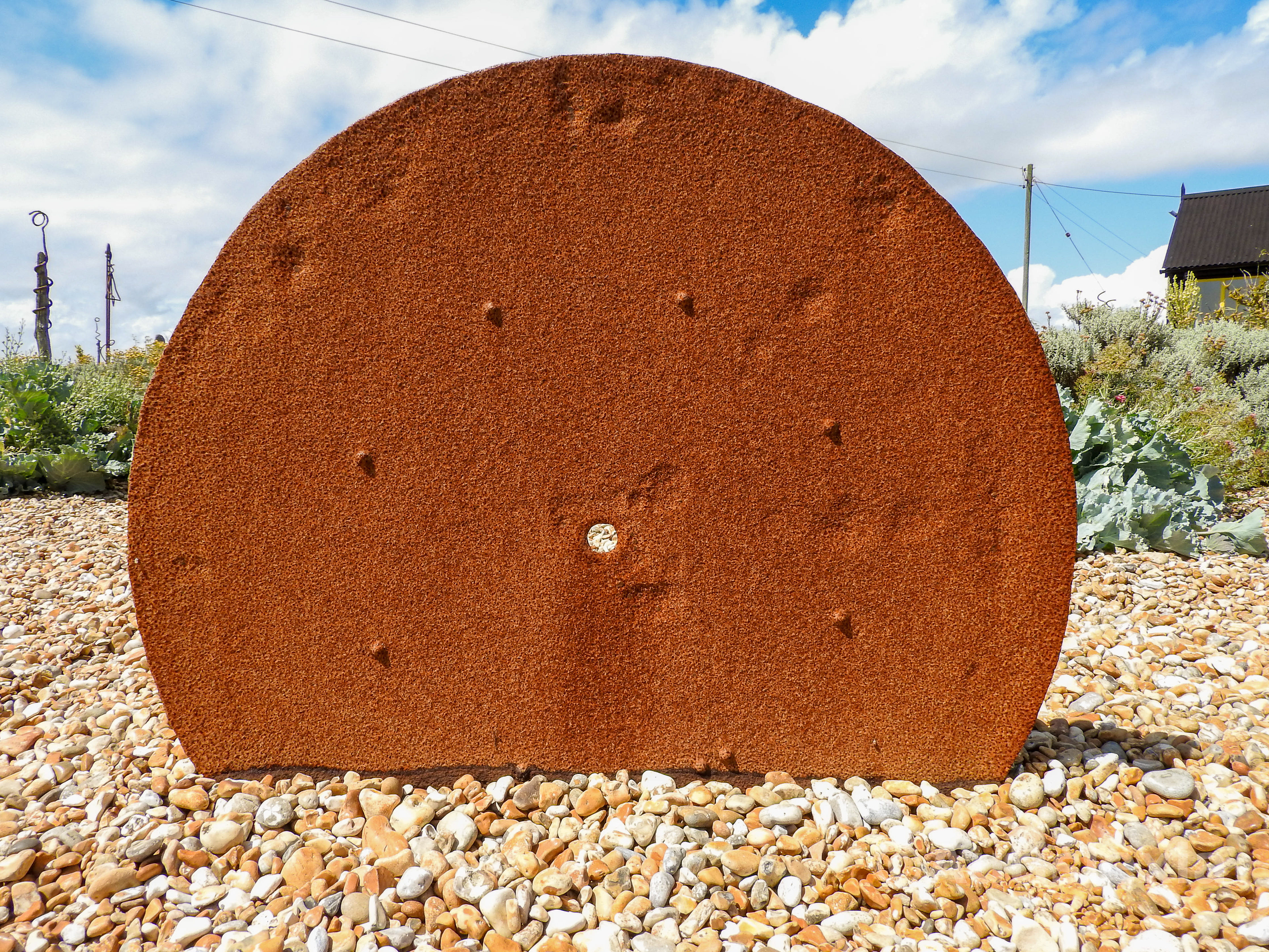 A rusty disc about three quarters sticking above the pebbles, it has a small hole in the middle and around that several rivets.