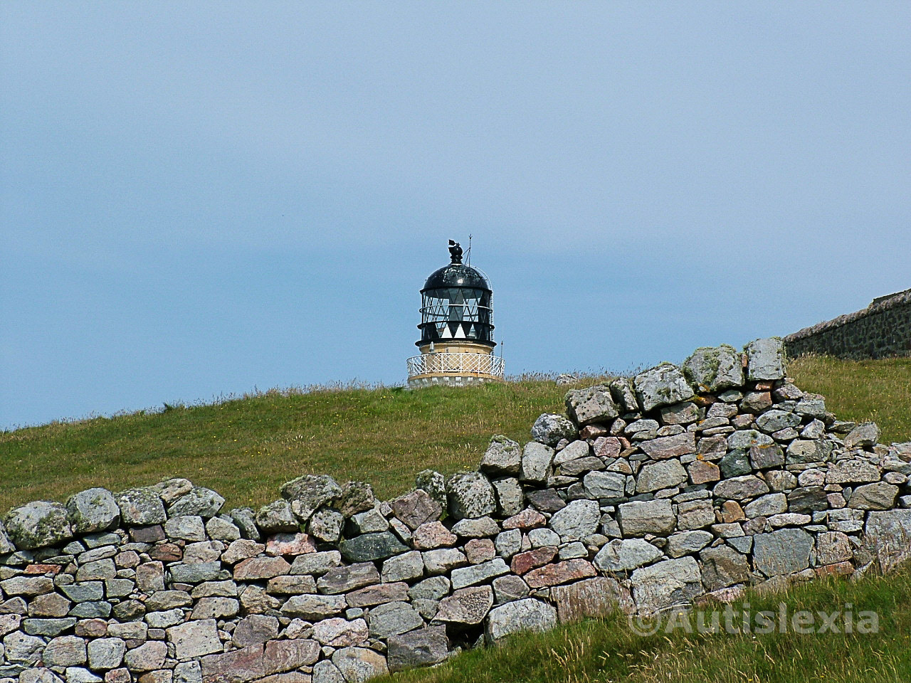 The top of Cape Wrath Lighthouse appearing out of the grass with a stone wall in the foreground