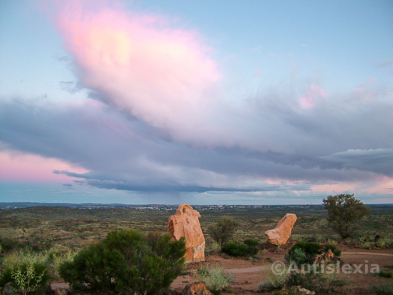 Image taken from the Living Desert Sanctuary and Sculpture Symposium, just a few miles outside Broken Hill, looking back at the town just before sunset, with pink tinged clouds dominating the image and rain falling in the distance.