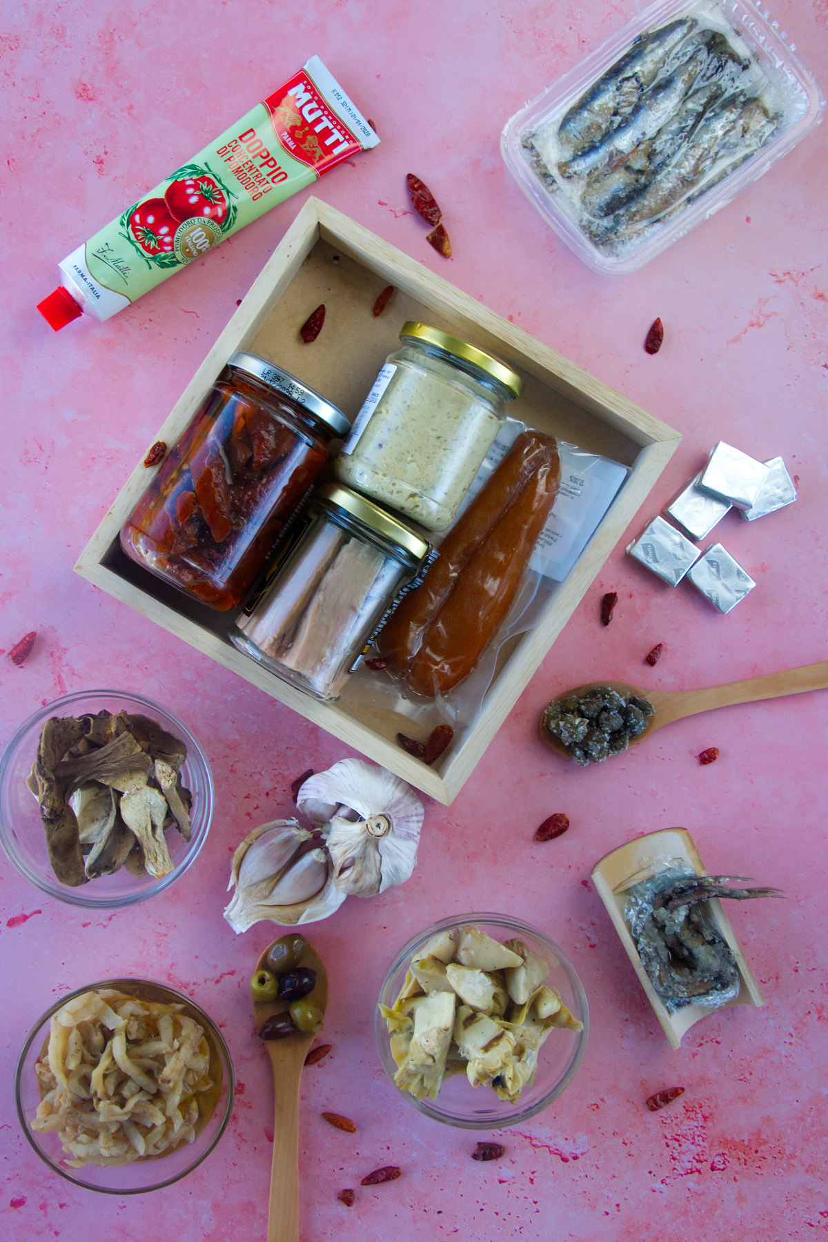 Vue de dessus : ingrédients du garde-manger italien sur fond rose avec tube de concentré de tomate Mutti, bocaux de tomates séchées et conserves, gousses d'ail et petits bols d'artichauts, champignons et olives.