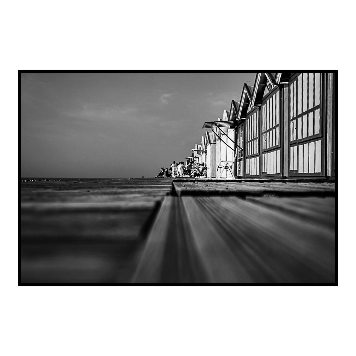 12. Déambulations sur les planches (août 2012) … trouver la bonne perspective. Cela fait des jours que je parcours les chemins de planches qui bordent les cabines de plage à CAYEUX-SUR-MER. J’y ai déjà pris des dizaines de clichés avec le tout nouvel appareil que je viens de me payer, sans véritablement trouver le moyen d’immortaliser l’endroit autrement qu’en mode « carte postale ». Je n’arrive pas à dégager de point de vue intéressant de mes nombreux allers et venues. Je veux que mon image soit une véritable scène de vie tout en offrant un regard non conventionnel sur ce qu’elle représente. Rien n’y fait, je ne trouve pas l’inspiration. Agacé, je prends la décision de m’asseoir un instant avant de repartir sans la moindre photo ce jour-là. Je tourne la tête et ai en ligne de mire cette famille elle aussi assise et qui profite du soleil. La perspective est intéressante et, sans véritablement réfléchir, je tente le tout pour le tout et m’allonge sur les planches. Je cadre rapidement avant d’activer le déclencheur et attends que le résultat s’affiche sur l’écran. Pas mal ! La profondeur de champ est un peu juste, mais l’angle est bon. Quelques réglages plus tard, je réalise enfin le cliché qui me convient et rentre enfin satisfait de ma virée de bord de mer.