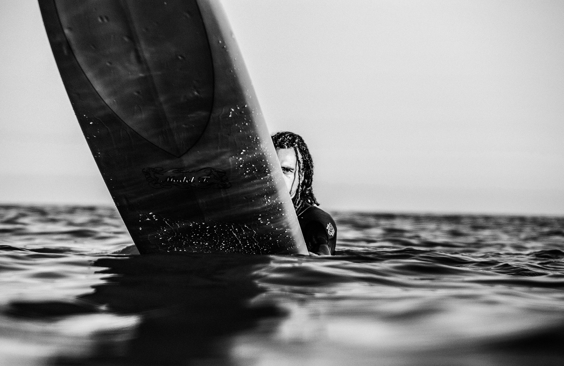 Fotografía de un surfista flotando en el agua y con media cara tapada por la tabla.