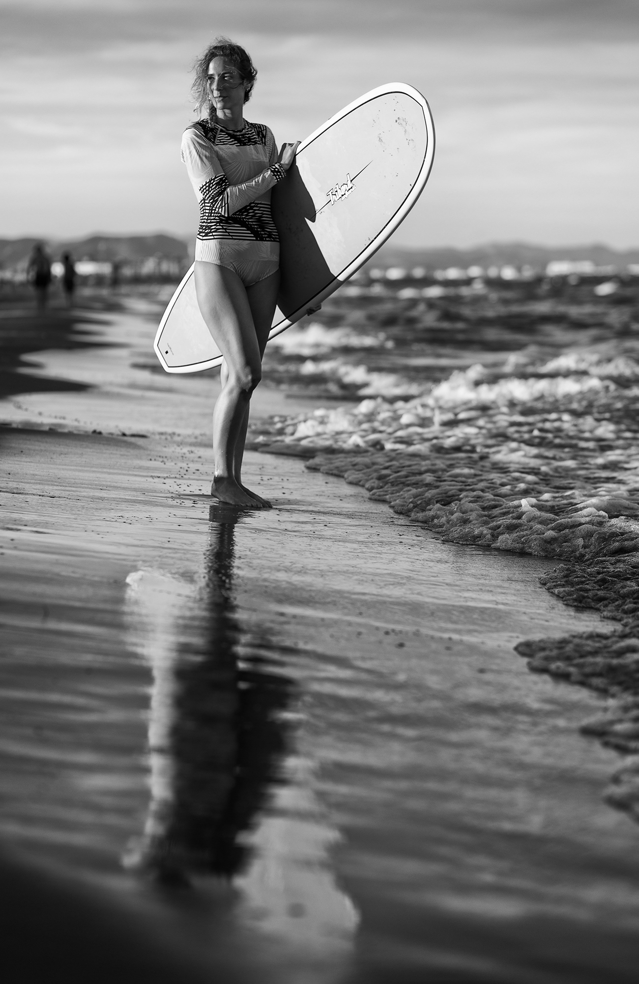 Fotografía en blanco y negro de una surfista de pie en la orilla de la playa y sujetando la tabla debajo del brazo.
