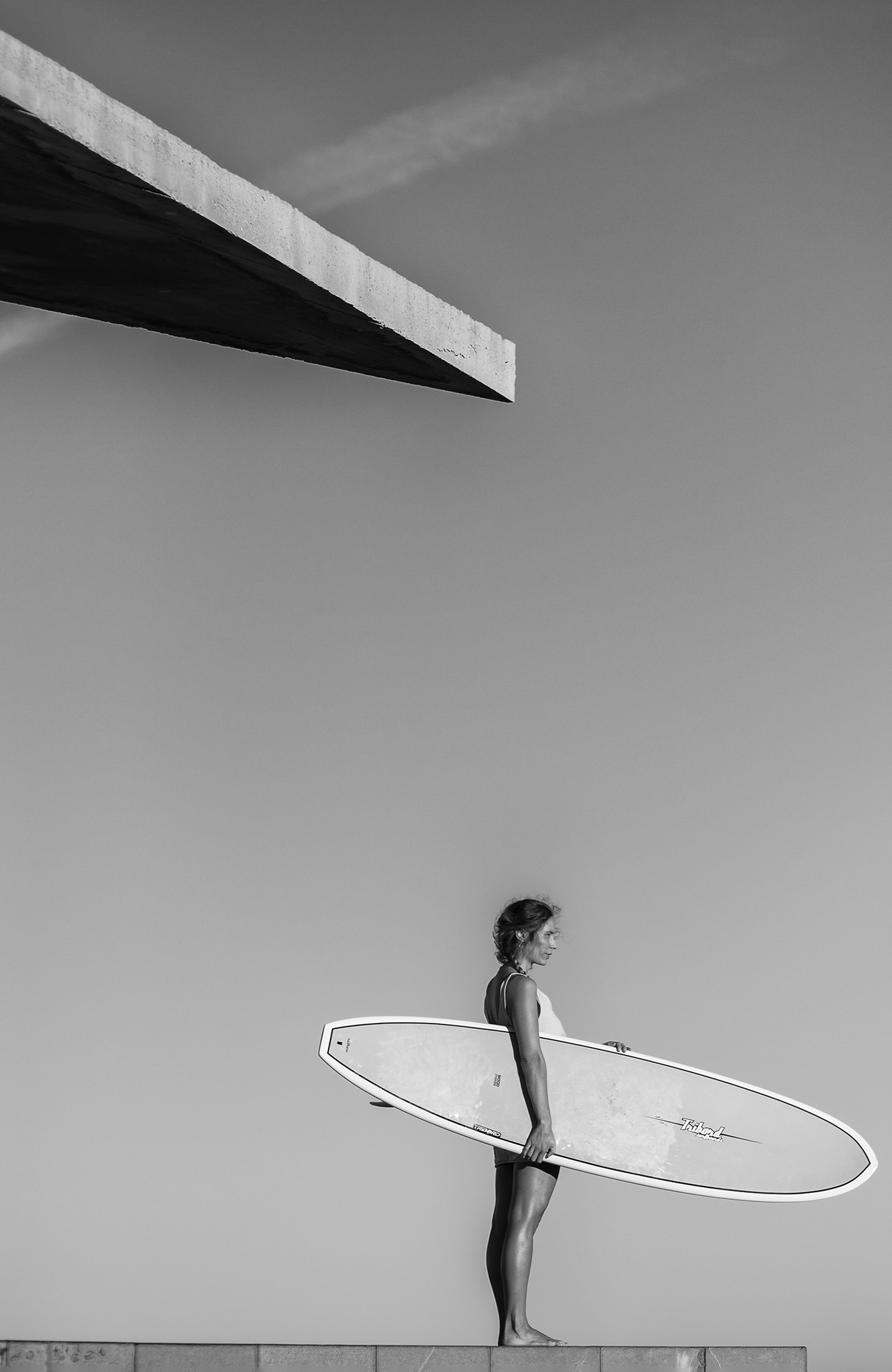 fotografía en blanco y negro de una surfista de pie con una tabla  bajo una cornisa de un edificio