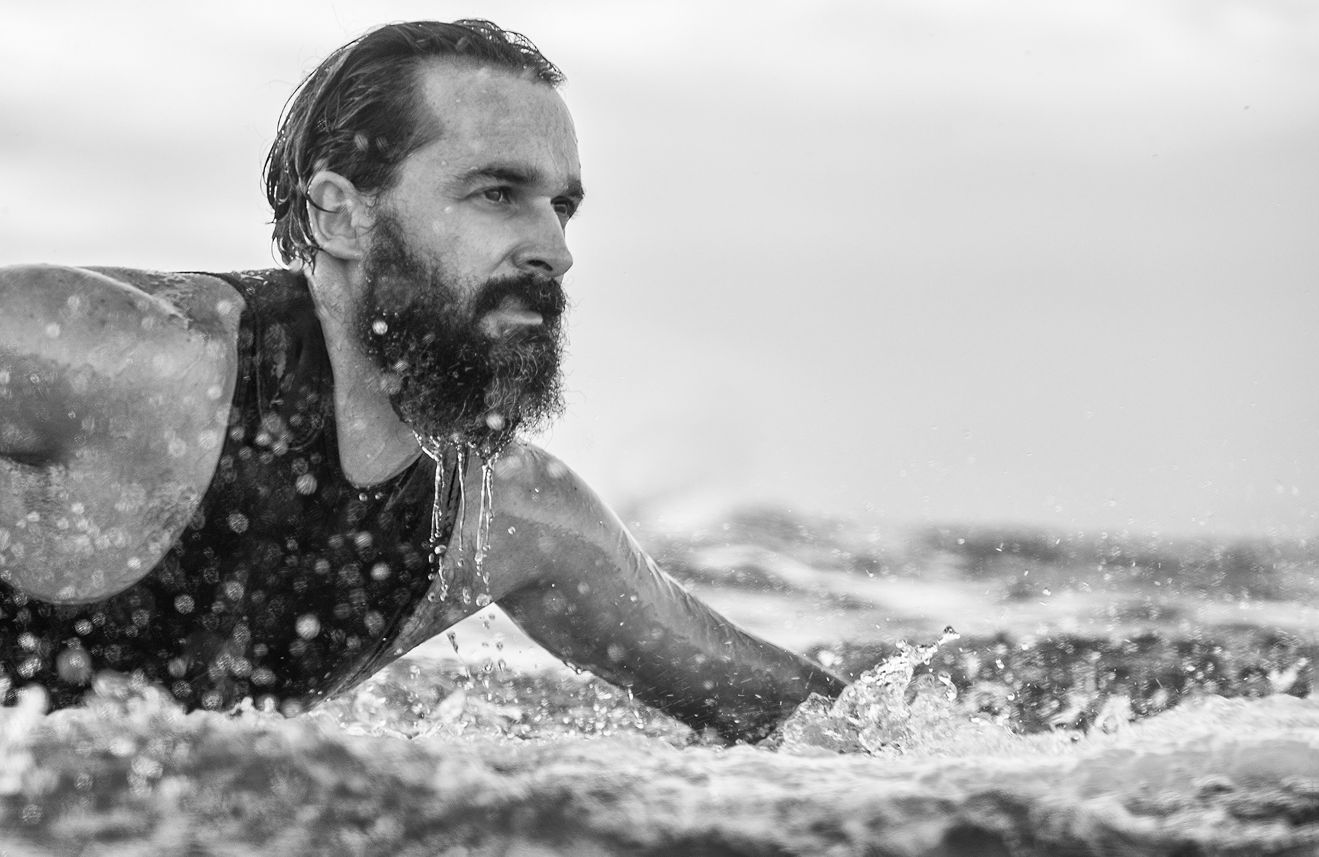 Fotografía en blanco y negro de un surfista de medio cuerpo, remando en el agua y con salpicaduras de agua por la barba