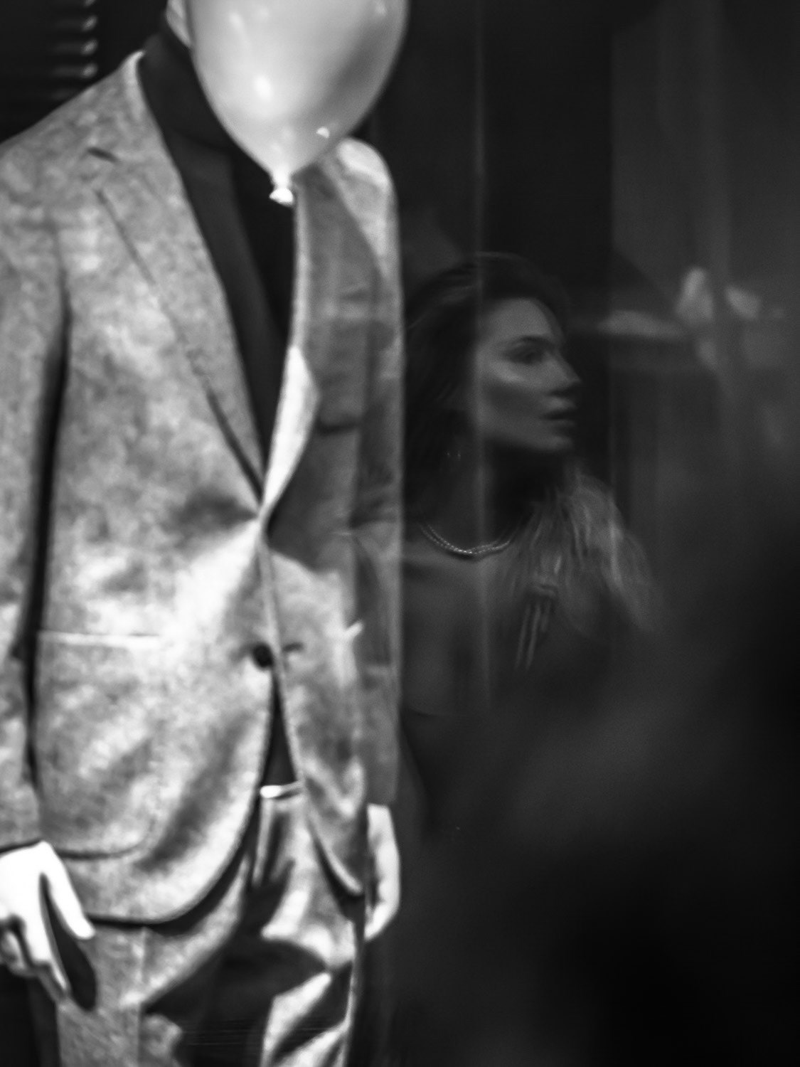 A woman’s reflection intertwines with a mannequin's balloon-shaped head, blurring the line between reality and artifice. The surreal juxtaposition evokes a film noir atmosphere, hinting at themes of hidden identities and societal masks. The mannequin lingers behind the balloon, transforming the moment into an uncanny tableau — a fleeting glimpse into a world where personas are crafted, concealed, and reflected back at us in fragments of truth.
