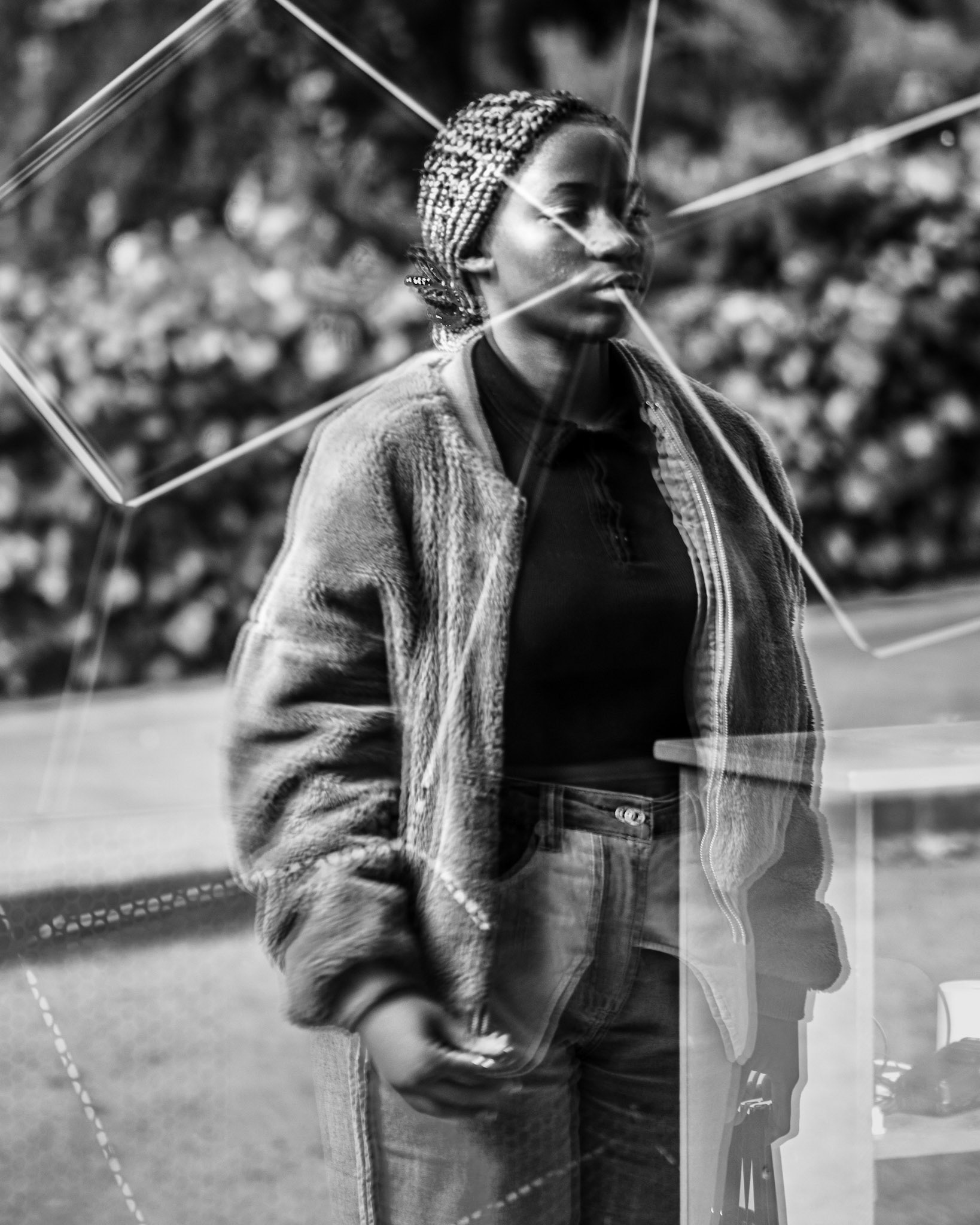 A woman walks with quiet strength, her reflection intersected by sharp geometric lines that seem to cut through her form. The fragmented world around her points directly at her, as if the universe itself is framing her presence, both challenging and elevating her identity. The glass fractures her essence, hinting at ancestral stories and unseen burdens, yet she remains resolute, embodying both resilience and transformation.