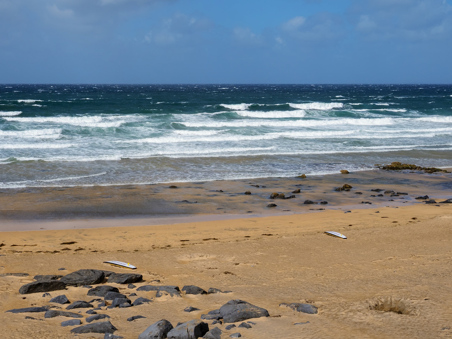 Das Bild zeigt eine Küstenlandschaft mit einem breiten Sandstrand und dem offenen Meer. Die Wellen brechen am Strand und erzeugen eine dynamische Szene. Der Himmel ist bewölkt, was eine eher düstere Stimmung erzeugt. Die Farben sind gedämpft, mit einem Schwerpunkt auf Braun-, Grau- und Blautöne. Die Perspektive ist weitwinkelig, was die Weite der Landschaft betont. Es wirkt wie ein typischer Tag an der Küste, vielleicht mit etwas rauem Wetter.